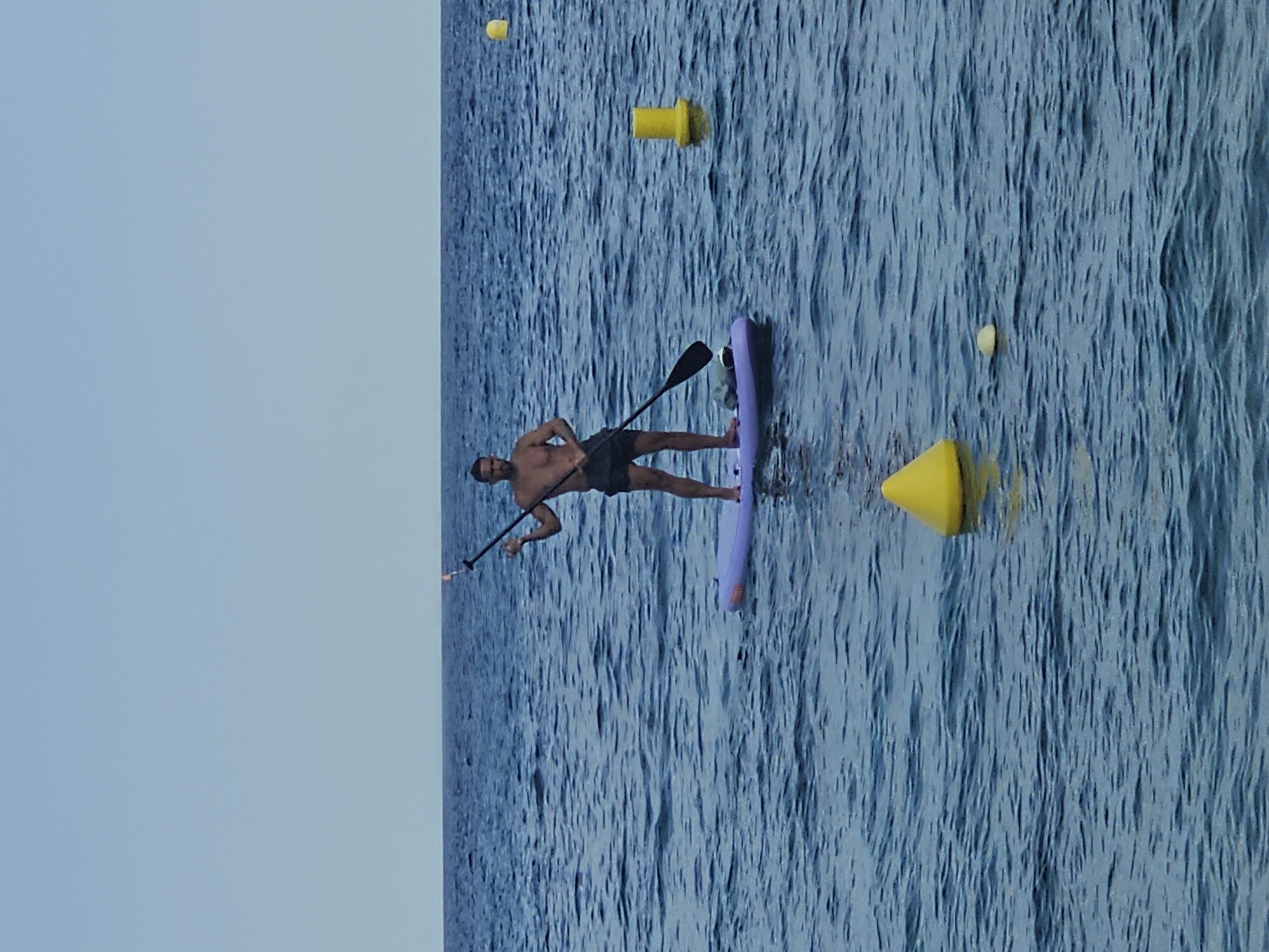 This image features a person stand-up paddleboarding on a body of water. The subject, a bare-chested man wearing dark shorts, is positioned on a light purple paddleboard and holds a paddle. The water is a deep blue with visible ripples, suggesting some choppiness or a gentle breeze. Several yellow buoys are scattered across the water, with one larger, cone-shaped buoy closer to the foreground. The background on the left is a clear, light blue sky, indicating a bright, likely sunny day. The orientation of the image suggests it was taken with the camera held vertically. The overall scene conveys a sense of outdoor recreation and activity on the water. The location is identified as Blanes, Spain, implying a coastal or lakeside setting. There is no visible text in the image.