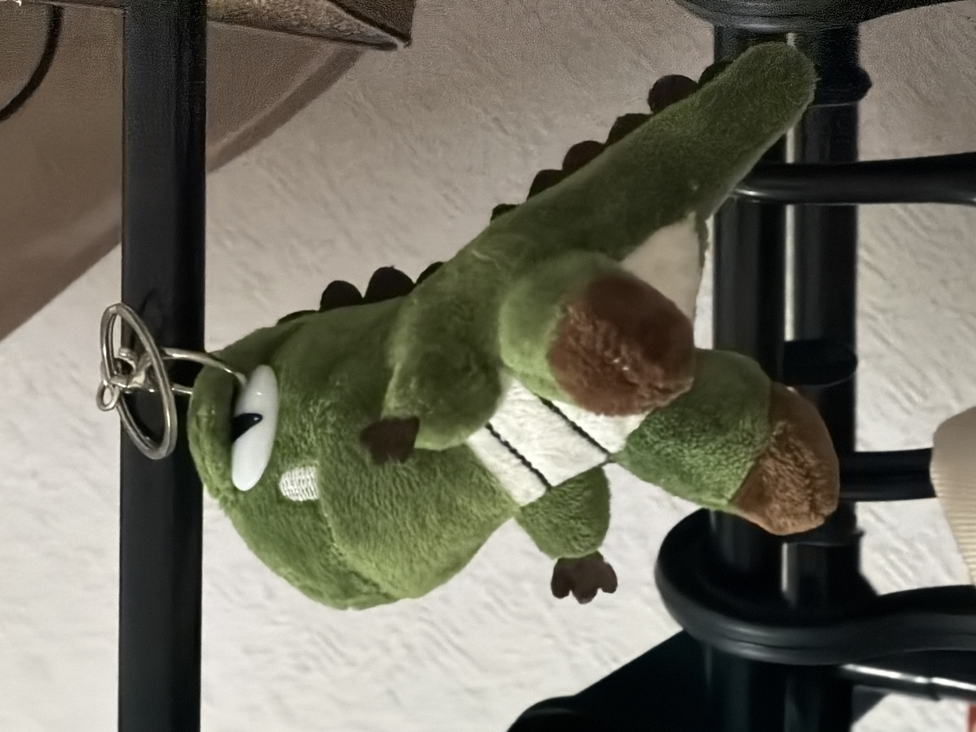 A green plush dinosaur keychain hangs from a black metal rod, possibly part of a closet or shelf. The dinosaur has white and brown accents on its body and feet, and a black eye with a white outline. The background is a textured, light-colored wall. The scene appears to be indoors, within a residential setting. There are no people or animals visible. There is no information to suggest this image was taken in Moscow, Russia.