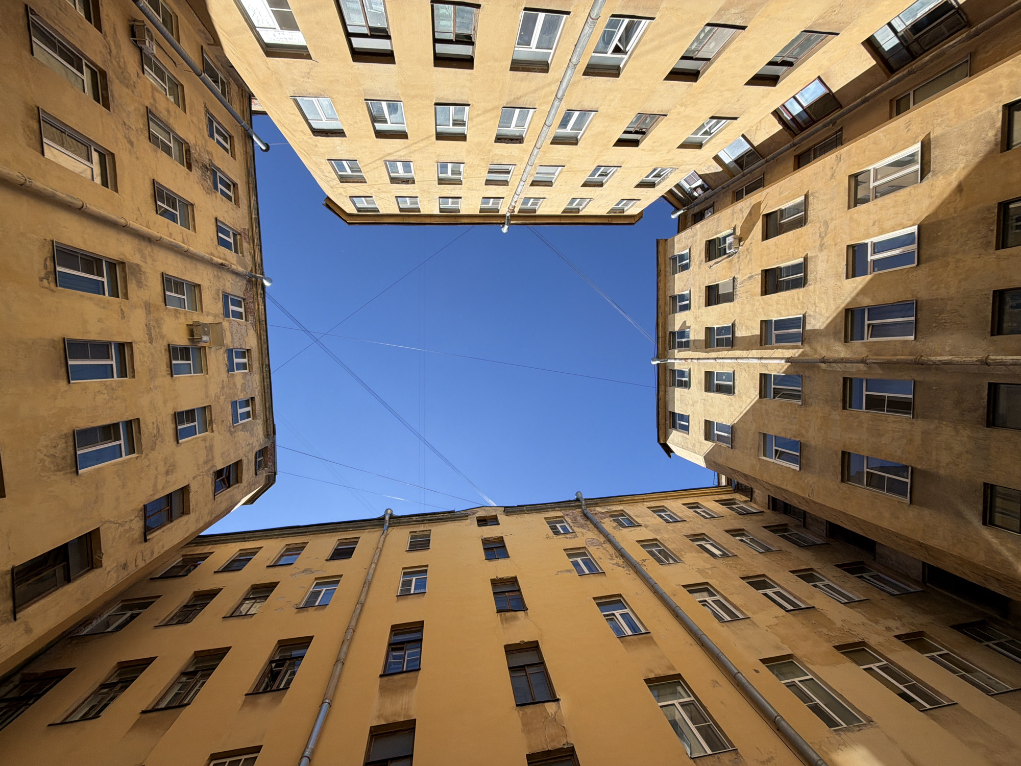 This image captures a view looking directly upwards into a narrow courtyard between tall, old, yellow buildings in Saint Petersburg, Russia. The architecture suggests a typical St. Petersburg courtyard, often referred to as "колодцы" (wells). The buildings have multiple stories with numerous windows, many of which are open or appear weathered. The facades are textured and show signs of age and wear. The sky above is a clear, bright blue, crisscrossed by thin electrical wires, a common sight in older city areas. The perspective is distorted due to the wide-angle lens and the upward angle, making the buildings appear to lean inwards and converge towards the sky. There are no people visible in the frame. The overall impression is one of urban density and historical architecture, emphasizing the unique spatial characteristics of St. Petersburg's courtyards.