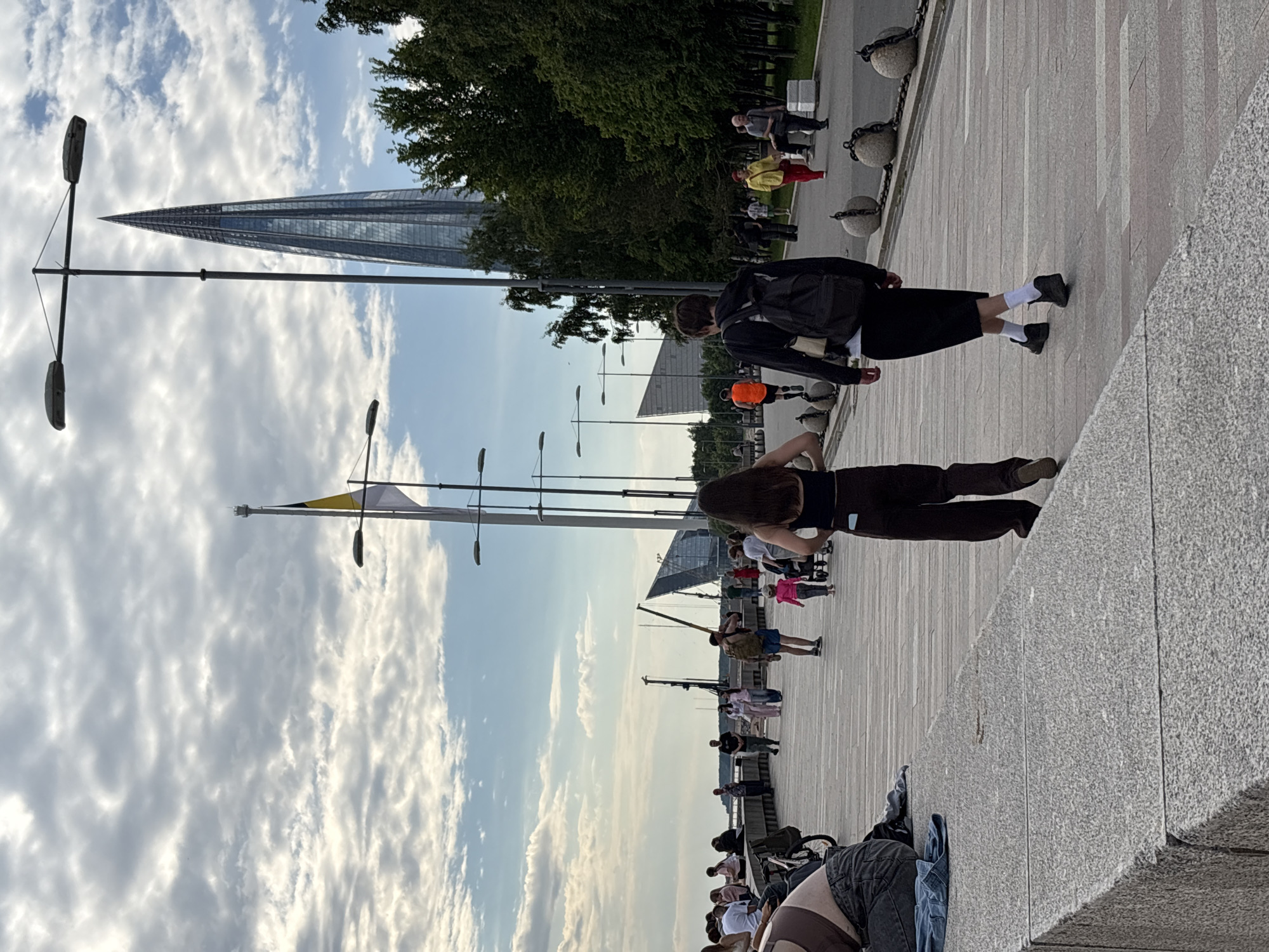 The image captures a vibrant urban promenade in Saint Petersburg, Russia, under a bright, partly cloudy sky. The perspective is slightly elevated, looking along a wide, paved walkway towards the horizon and a distinctive modern skyscraper.
In the foreground, a person with long dark hair, clad in a dark top and brown trousers, is seen from behind, leaning slightly forward. Adjacent to them, another individual walks away from the viewer, wearing a black jacket, black skirt, white socks, and dark shoes, carrying a black backpack. The ground is a light grey paved surface, and to the right, a textured grey granite ledge or wall defines the edge.
The mid-ground is populated with numerous people engaged in leisurely activities like walking and strolling. Notable figures include one person in a bright yellow top and red trousers, and another in an orange top. Concrete bollards connected by chains delineate the right side of the promenade, bordering a green area with trees. Beyond the trees, other modern buildings are visible.
The prominent Lakhta Center skyscraper, with its iconic pointed, glass-paneled design, stands tall on the left side of the background, reflecting the sky. Other contemporary glass structures are also discernible further right. Several street lamp posts with dual light fixtures and a flagpole displaying a yellow and white flag are spaced along the promenade.
The weather is bright and pleasant, with a clear blue sky dotted with fluffy white and grey cumulus clouds. The soft lighting suggests it is daytime, possibly late afternoon. The scene depicts a bustling public space, emphasizing modern architecture and urban leisure. No visible text is present.