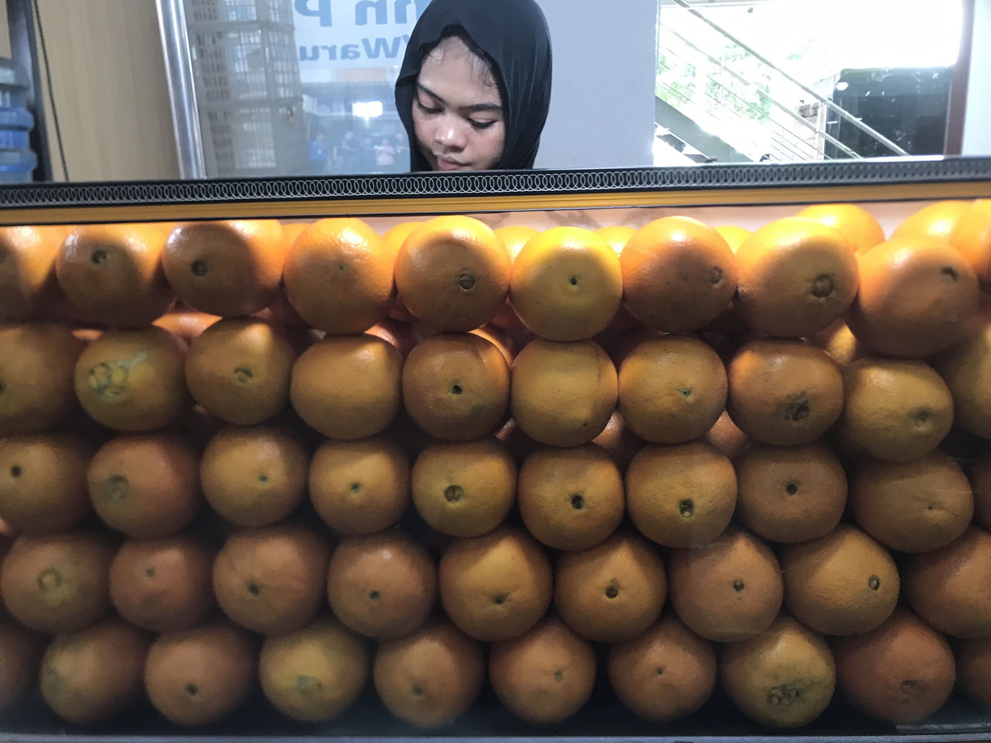 A young woman with a dark headscarf stands behind a display case filled with stacked oranges. The oranges are arranged in a grid pattern and appear to be the focus of the shot. The location is indicated as City of Medan, Indonesia.
