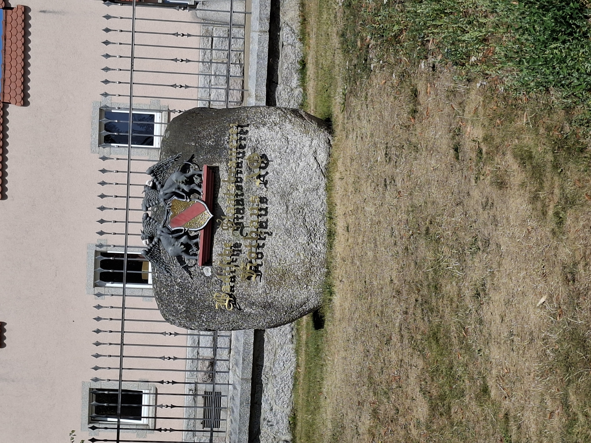 A weathered stone monument bearing an inscription and a coat of arms is situated in front of a light-pink building with a red-tiled roof. The monument is partially obscured by a black metal fence. The inscription on the stone appears to be in German and is difficult to fully decipher, but it may be related to the town hall or a historical event. The coat of arms features two winged mythical creatures supporting a shield with red and yellow elements. The ground in front of the monument is covered with dry grass and some scattered weeds. The building behind the fence has two rectangular windows with decorative stone surrounds. The overall scene suggests an outdoor historical marker in a public space.