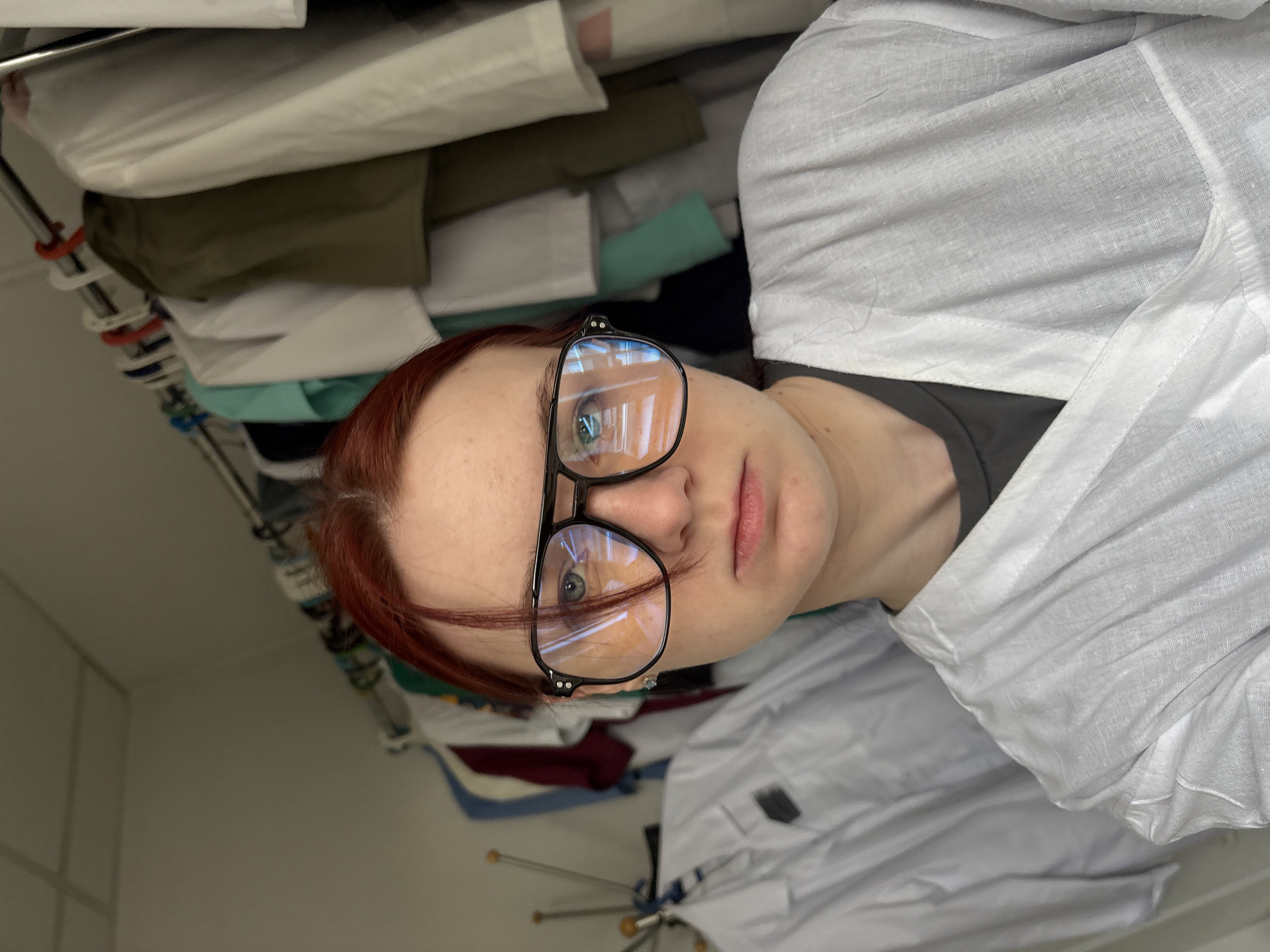 The image shows a young woman with red hair and glasses, looking directly at the camera. She is wearing a white smock over a dark t-shirt. In the background, there is a rack of clothing, possibly lab coats or uniforms, hanging on a rod. The lighting suggests an indoor setting. There is no visible indication of the location being in Saint Petersburg, Russia.