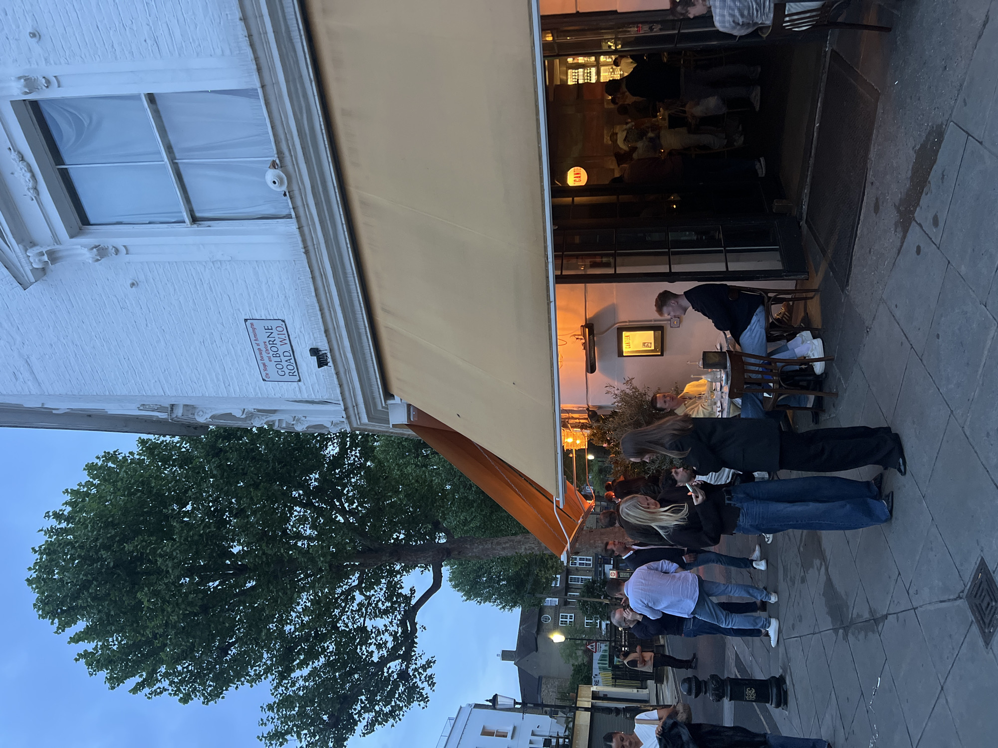 Here is a description of the image:
This is an outdoor scene on Golborne Road, located in the Royal Borough of Kensington and Chelsea, London, United Kingdom. A group of people are congregated on the sidewalk outside a building with a large awning. Some appear to be waiting in line. There is outdoor seating beneath the awning in front of the building, and people are seated at the tables. One person is standing by a table and seems to be interacting with a device. The building has white brick and a window with a light-colored covering inside. A large tree is visible to the left. The restaurant seems to be named "Canteen" given the sign at the entrance.
