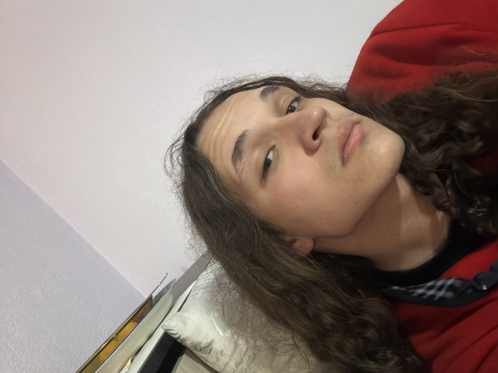 A person with long, wavy brown hair is looking at the camera. They are lying down on a pillow. They are wearing a black shirt and a red hoodie. The background is a white wall. The location is unidentifiable as Asenovgrad, Bulgaria.