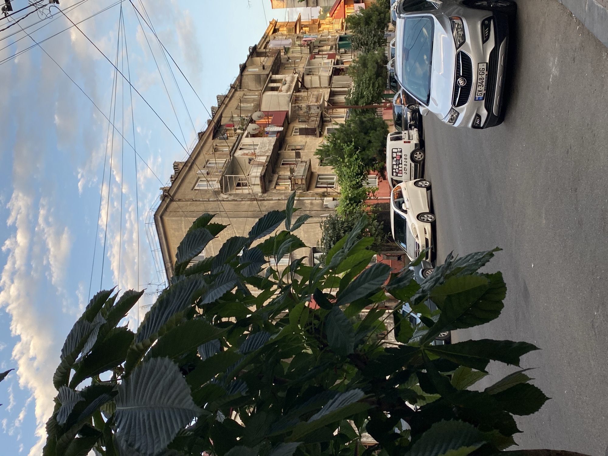 The image depicts a street scene in Batumi, Georgia, featuring a multi-story apartment building on the right and a large tree with dark green leaves in the foreground on the left. The building is weathered and appears to be from a Soviet-era construction, with numerous balconies, some enclosed with glass or metal, and clothes hanging from some of them. Several cars are parked along the street, including a silver Buick SUV in the foreground with license plate GX-348-GG. A white van with writing on its side is parked behind a white car. The sky is blue with scattered white clouds, and electrical wires crisscross overhead. The overall environment suggests an urban residential area.