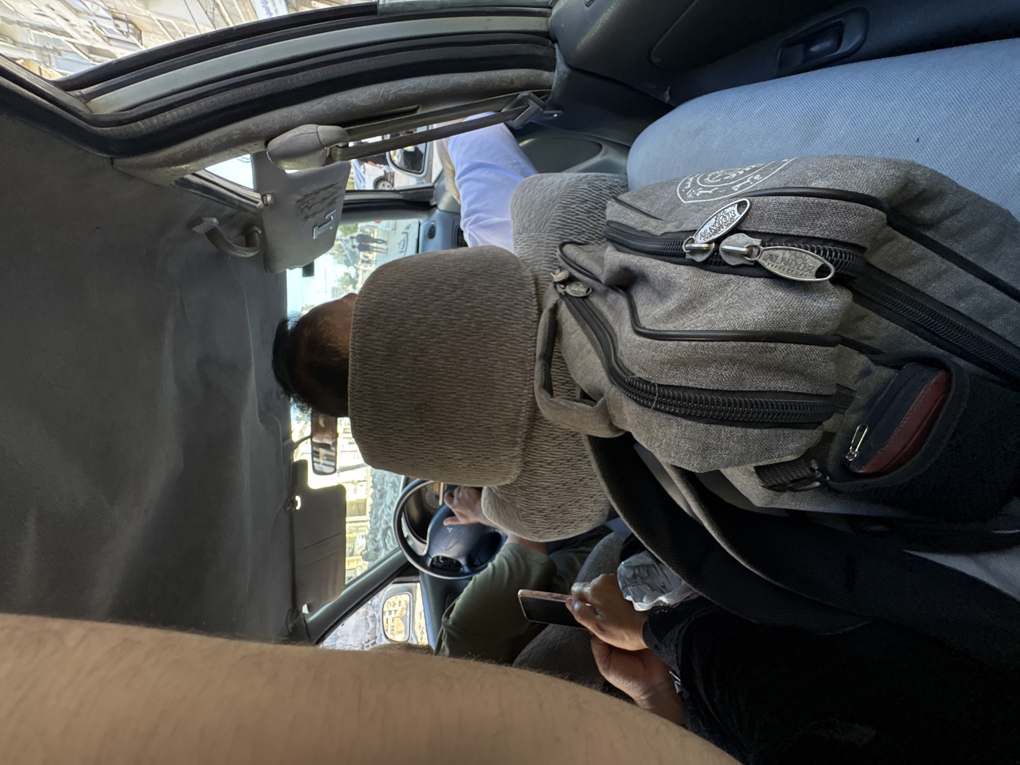 In the interior of a vehicle, a person is seen driving, with their hands on the steering wheel. The vehicle appears to be in motion, with the surroundings visible through the windows. A gray backpack is placed on the passenger seat, with zippers that have the text "ALNOOR" visible on them. Another person is seated in the vehicle, holding a smartphone. The scene is illuminated by natural daylight, suggesting it is daytime. The setting is consistent with an urban or suburban environment, possibly in Gaza, Palestinian Territories, given the provided location context. The individuals' facial expressions are not clearly discernible.