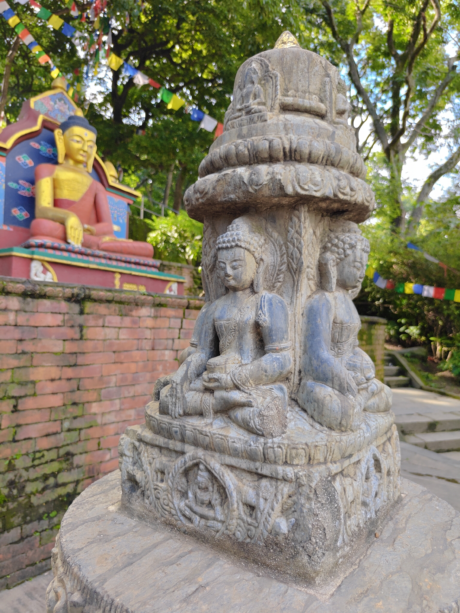 This image captures a stone carving of four Buddha statues in a seated meditative pose, arranged around a central pillar. The statues are weathered and detailed, showing intricate folds in their robes and serene facial expressions. The pillar itself is adorned with carvings and topped with a dome-like structure, possibly representing a stupa.
In the background, a larger, brightly colored Buddha statue is visible, seated on a raised platform with intricate decorative elements. This statue is predominantly gold and red. The scene is set outdoors, amidst lush green trees. Colorful prayer flags are strung across the branches of the trees, adding a vibrant element to the otherwise earthy tones of the stone carvings and brick wall. The ground appears to be paved with stone.
The presence of prayer flags and the style of Buddhist iconography strongly suggest the location is within the Kathmandu Metropolitan City, Nepal, likely at a Buddhist monastery, temple complex, or stupa site such as Swayambhunath or Boudhanath. The brick wall to the left indicates a man-made structure, possibly part of the surrounding architecture. The lighting suggests it is daytime, with sunlight filtering through the leaves of the trees, creating dappled patterns on the ground.