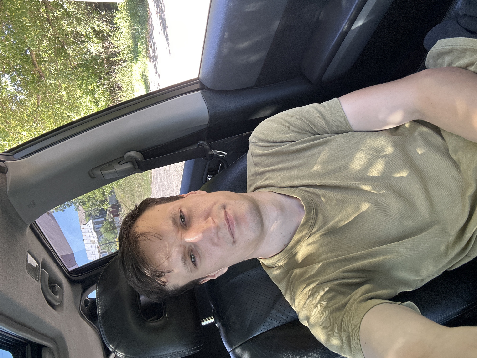 A man with brown hair and light skin, wearing a khaki t-shirt, is seated in the driver's seat of a car. The car's interior is visible, including the black leather seats and part of the dashboard and headliner. Sunlight streams in from the right side of the vehicle, casting shadows on the man's face and clothing. Through the car window, a building with a white roof and trees can be seen, suggesting an outdoor location. The image appears to be a selfie taken by the man. The scene is set in Makiv, Ukraine.