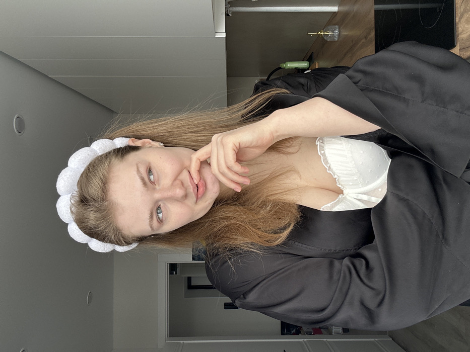 A single female subject is depicted in an indoor environment in Kazan, Russia, likely a modern kitchen or living area. She possesses light skin and long, light brown hair, styled with a white, puffy headband. She is dressed in a white, polka-dotted bralette or bustier visible beneath an open black satin robe. Her left hand is raised, with the index finger making contact with her lower lip. Her head is slightly tilted, and her eyes are directed towards the upper left of the frame, away from the camera. The background on the left features a doorway leading into a darker room and a light-colored wall. On the right, a light-colored wooden countertop is visible, hosting a black induction stovetop, a green container, and a small, clear glass object with a gold-colored top. Above the counter, light grey kitchen cabinets are installed. The scene is evenly lit, suggesting natural light from an unseen source to the left.