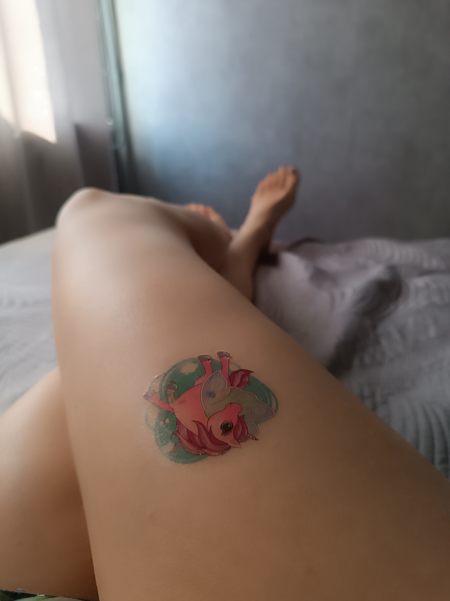 A person is lying on a bed with their legs crossed. A sticker of a cartoon unicorn is on their upper thigh. The setting is an interior with a window to the left.