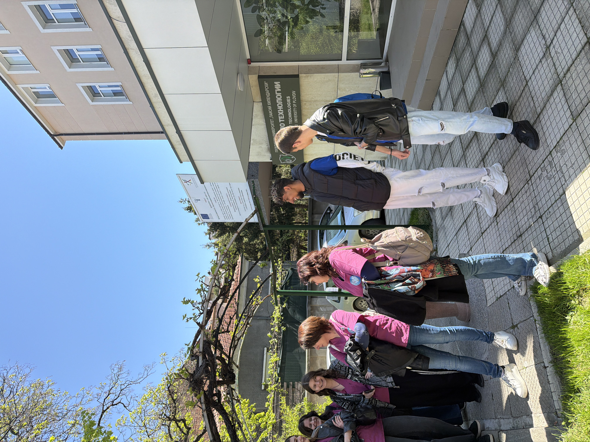 The image captures a group of people gathered outdoors on a bright, sunny day in Plovdiv, Bulgaria, in front of a university building.
On the left side of the frame, a group of approximately six women and girls is visible. Most are dressed in bright pink or magenta tops, dark jackets, and jeans, suggesting a uniform or shared affiliation. They are carrying various bags and some are smiling. They stand on a paved area, with an overhead arbor covered in budding green vines and trees with fresh spring foliage providing natural shade.
To the right, two young men stand near the entrance of a building. One wears a black leather jacket, light ripped jeans, and a backpack, while the other is in a black puffer vest over a white top, white ripped jeans, and a backpack.
The main building in the background features both traditional architecture with rectangular windows and a modern section with large reflective windows. A prominent sign on this building displays text in both Cyrillic and English, clearly reading "ПЛОВДИВСКИ УНИВЕРСИТЕТ „ПАИСИЙ ХИЛЕНДАРСКИ“" (Plovdiv University "Paisii Hilendarski") and "UNIVERSITY OF PLOVDIV," with "TECHNOLOGIES" also visible, likely indicating a faculty like Information Technologies. A white sign with the European Union logo and another logo featuring a red 'X' is visible further left, possibly detailing a project.
The sky is a clear blue, and the strong shadows cast by the people and buildings suggest it is daytime, likely mid-morning or early afternoon. The overall scene depicts a casual gathering, possibly students or participants in an event, outside a university facility.
