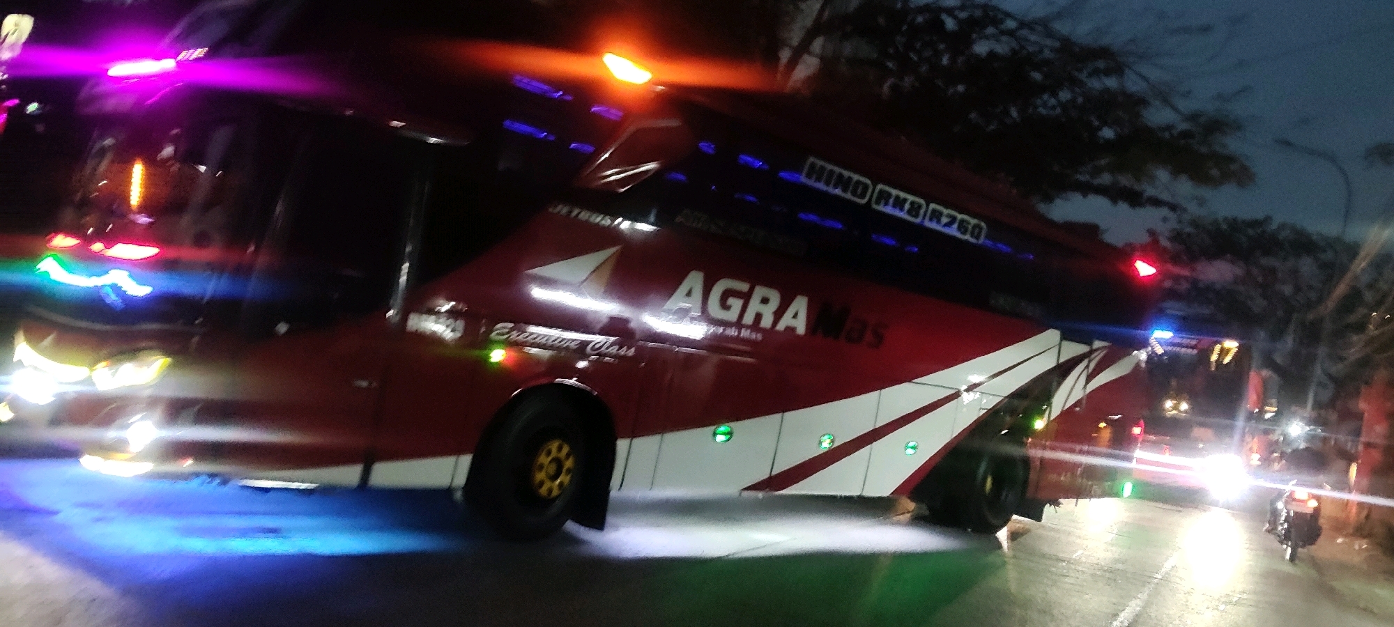 The image shows a red and white bus labeled "AGRA MAS" and "JETBUS" parked on a street at night. The bus has a sleek design with a prominent logo and text indicating it is an "Executive Class" vehicle. Various lights, including neon and flashing lights, illuminate the bus and the surrounding environment. The street appears wet, suggesting recent rain, and the lighting casts reflections on the surface. Trees are visible in the background, and the sky is dark, indicating nighttime. There are a few blurry figures and vehicles in the distance, including what appears to be a motorcycle. The scene suggests a bus station or a roadside stop in a city, potentially Pasirsari, Indonesia, given the specific markings on the bus.