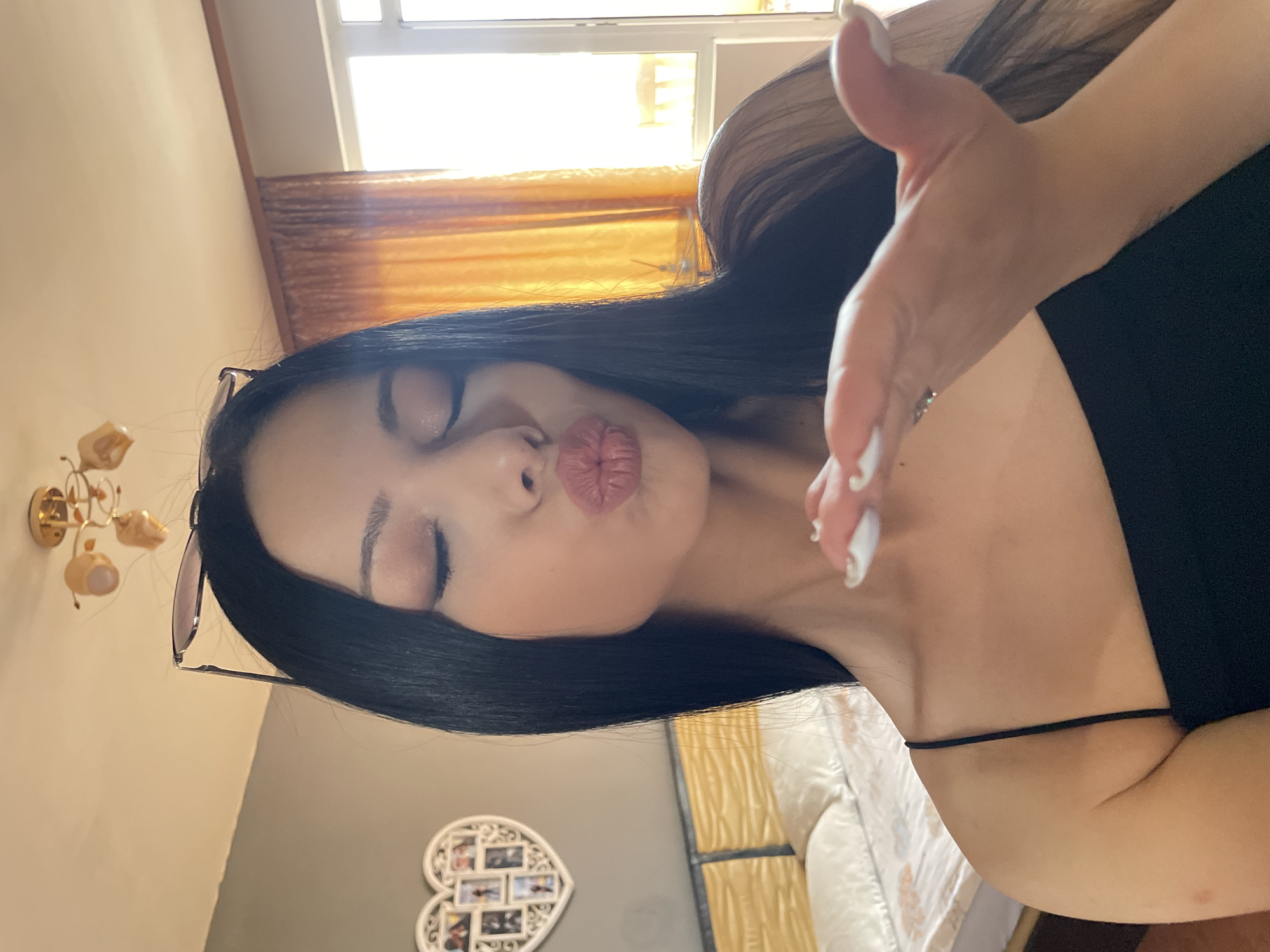 A young woman with long, dark hair and makeup is blowing a kiss towards the camera. She is wearing a black tank top. Her eyes are closed and her lips are pursed. She has sunglasses perched on her head. In the background, a bed with white linens is visible, as well as a heart-shaped picture frame on the wall containing several photographs. A chandelier hangs from the ceiling, and orange curtains cover a window. The scene appears to be indoors, likely in a residential setting, such as an apartment in Stara Zagora, Bulgaria.