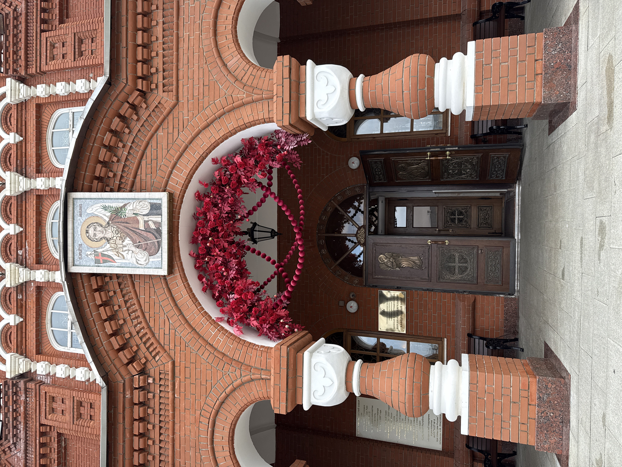 The image shows the entrance to a red brick building, likely a church or cathedral, decorated with red foliage and spherical ornaments. Above the entrance, a mosaic depicts an angelic figure holding a staff and a palm frond. The building features arched windows and ornate doors. To the left of the entrance is a plaque detailing the history of the building, and benches are placed along the exterior. The scene is set in Izhevsk, Russia, specifically at the Mikhailo-Arkhangelsky Cathedral. The overall impression is one of religious and historical significance, with festive decorations adorning the entrance.