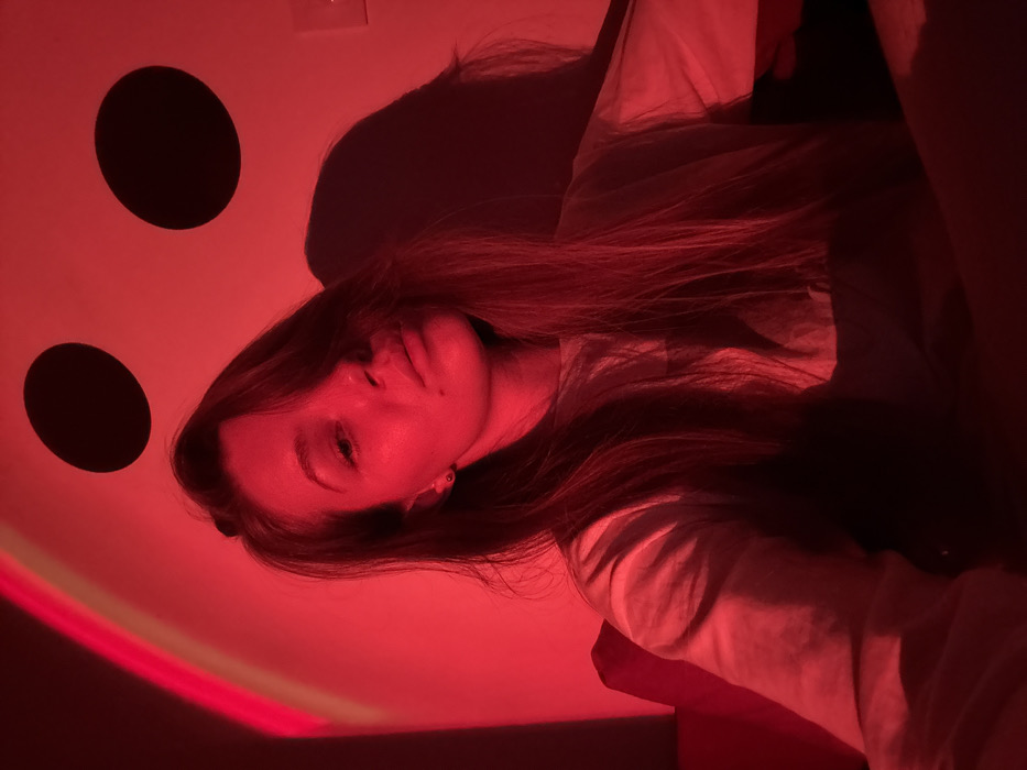 A female subject is depicted in a medium-shot, illuminated predominantly by a red light source, in an indoor setting within Vozdovac Urban Municipality, Serbia. The subject has long, dark brown hair, parted on the right and falling over her shoulders and chest. She looks directly at the camera with her mouth slightly ajar. A small blemish or mole is visible on her left cheek, and a stud earring is present in her left ear. She is wearing a light-colored, short-sleeved t-shirt. The background features a light-colored wall with a curved, lighter red light projection on the upper left. Two large, distinct circular black decals are affixed to the upper right wall, above her head. A wall-mounted electrical fixture is visible on the far right. A dark shadow is cast on the wall behind the subject's right shoulder, likely from her form.