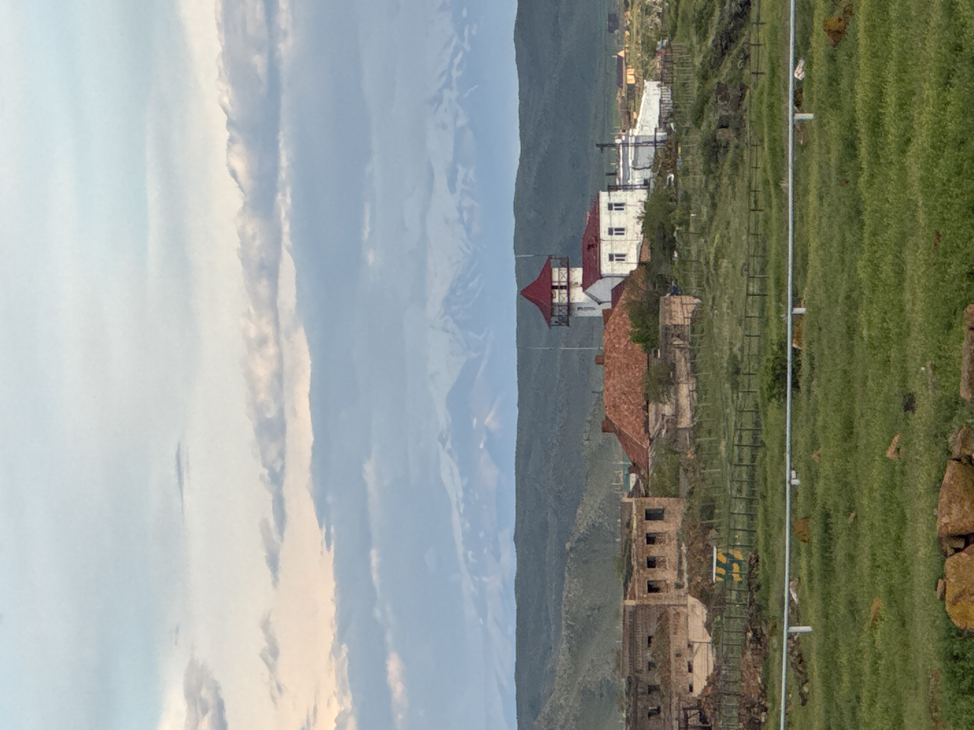 The image depicts a vertical view of the city of Anipemza, Armenia. On the right side of the frame is a field of green grass with a wire fence running along its edge. In the center, various buildings are visible, some of which appear to be partially ruined or under construction. A building with a red roof is prominent in the middle ground, while a white building stands slightly behind it. The background features a mountain range, against a sky filled with clouds.