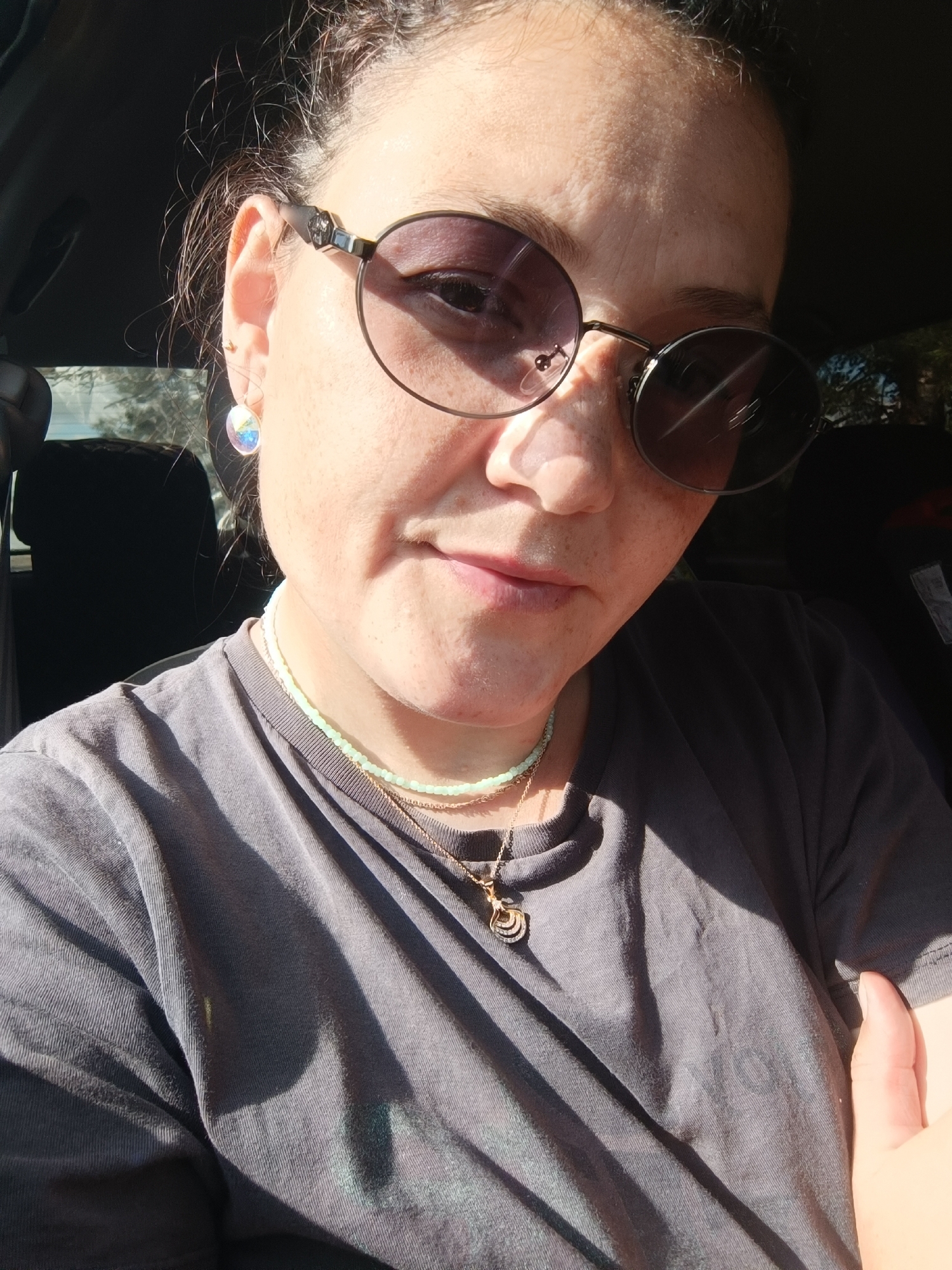 A woman wearing sunglasses and a grey t-shirt is pictured from the chest up. She has earrings and two necklaces on. The lighting suggests she is outdoors, possibly in a car, with sunlight filtering through the window. No specific location in Foki, Russia, can be determined from the image.