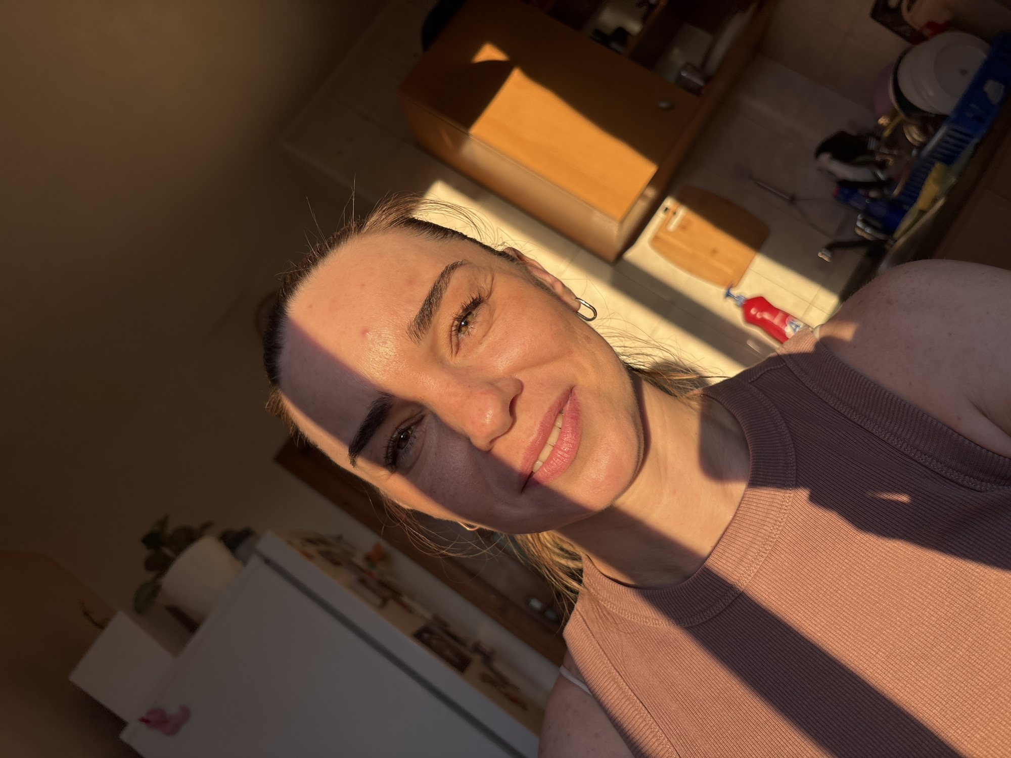 A close-up selfie of a woman with light skin, brown hair, and brown eyes, taken in a kitchen. She is wearing a pink ribbed tank top. The setting sun casts a warm glow and strong shadows across her face and the room. In the background, a refrigerator, wooden cabinets, a sink with dishes, and a cutting board are visible. The overall impression is casual and domestic.