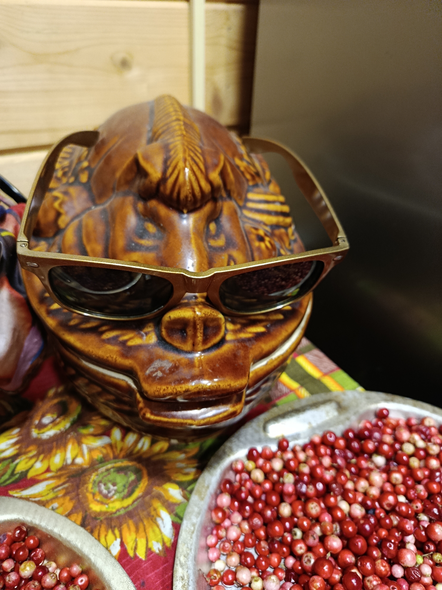 An overhead shot shows a brown ceramic bear head wearing gold-rimmed sunglasses. The bear head is decorated with vertical lines across the forehead, creating a textured effect. In front of the bear head, a tray of small, red berries is visible. Behind the bear head, a wooden plank wall can be seen. The bear head is placed on a colorful fabric that features a sunflower pattern. The scene appears to be indoors, possibly in a kitchen or a display area. There is no indication of the city of Верея, Russia, within the image itself.