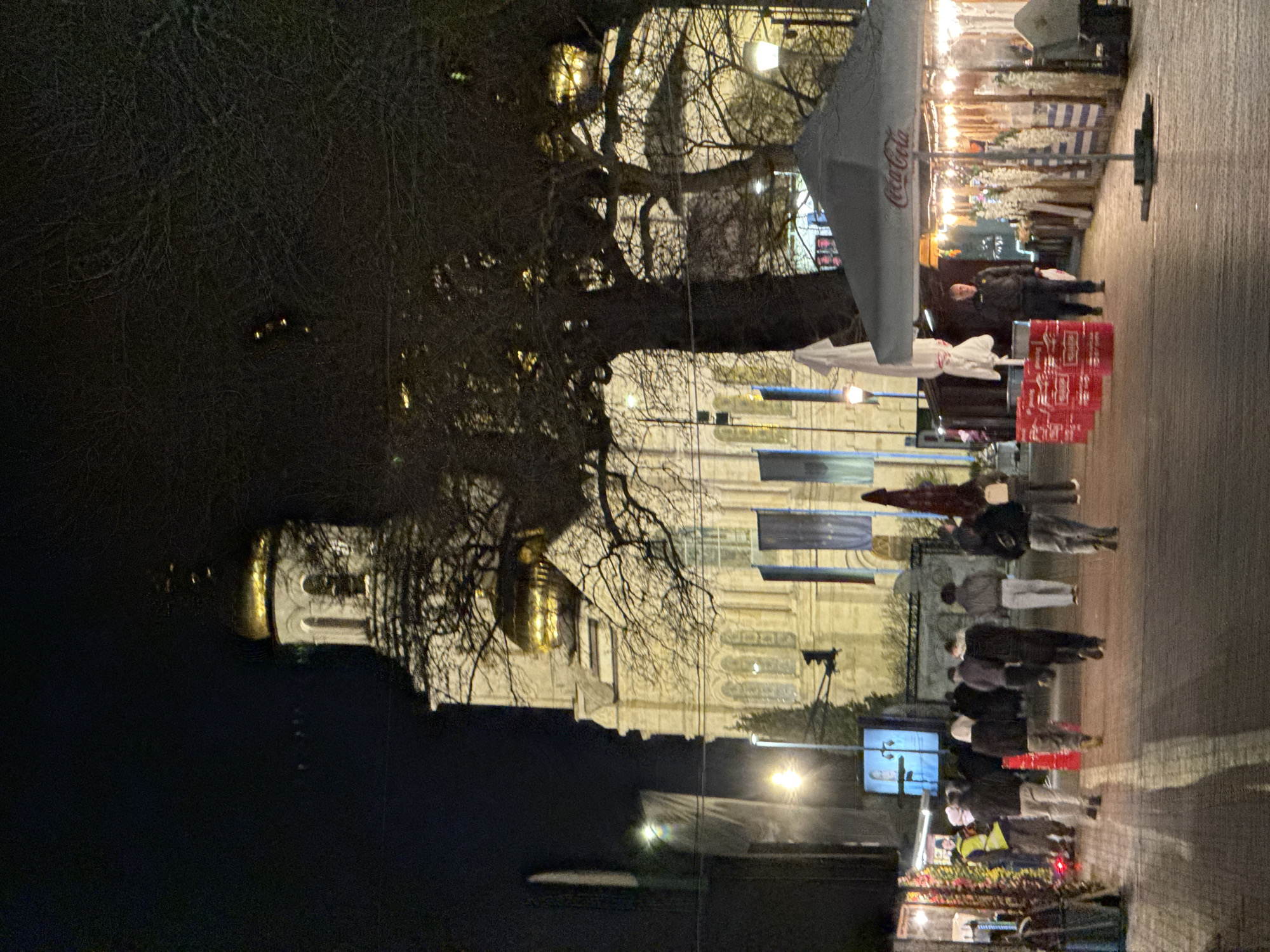 This is an outdoor nighttime scene showing a building with a golden dome in the distance, possibly a cathedral or church. The architectural style appears Eastern European. In the foreground, there is a bustling area with people walking along a paved surface. A Coca-Cola branded umbrella is visible, suggesting a vendor or stall. There are also flags or banners hanging overhead. The presence of people, vendors, and illuminated structures indicates a public space or marketplace in an urban setting. The overall environment is dark, with streetlights providing illumination. Given the architecture and the indication of the presence of flags/banners, it is possible this is taken somewhere in Europe, potentially the suggested area of Varna, Bulgaria.