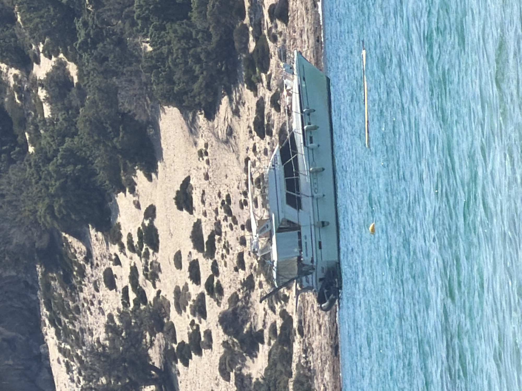 An eye-level, medium shot captures a light-colored motor yacht grounded on a rocky shoreline in the foreground. The boat is positioned parallel to the shore, facing left, with its stern closer to the viewer. The water in front of the boat is a vibrant turquoise, with gentle ripples reflecting the sunlight. A yellow buoy floats in the water to the left of the boat, and a long, narrow yellow object, possibly a raft or debris, is visible in the water to the right. The background consists of a sandy, arid hillside dotted with sparse green shrubs and trees, characteristic of a Mediterranean landscape. The sky is not visible.