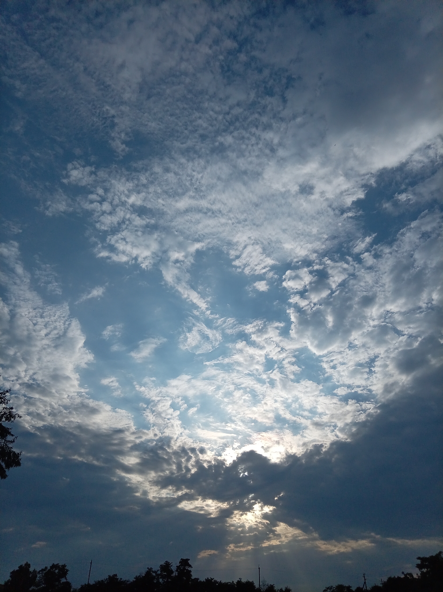 The image displays a sky filled with a mix of light and dark clouds, with the sun partially obscured by the darker formations. The clouds are varied in texture and density, with some appearing wispy and high-altitude, while others are thicker and lower. Sunlight is breaking through in certain areas, creating illuminated patches and highlighting the edges of the clouds. The bottom of the image shows a dark silhouette of trees and possibly some distant structures against the bright sky. The overall impression is of a dynamic sky, possibly near sunset or sunrise. There are no people or visible actions depicted. The location is specified as Гражданское, Russia.