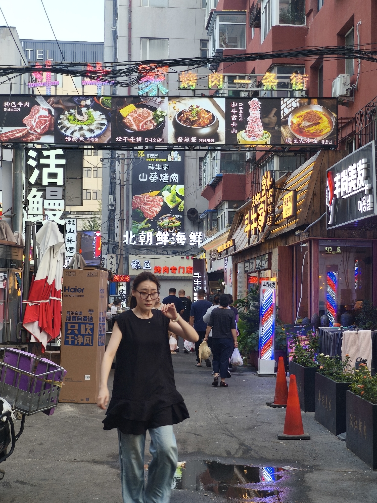 A street scene in Heping District, China, features a woman walking towards the foreground in a black top and light blue jeans, wearing glasses. She is looking to her right and her left hand is raised. Behind her, several people are walking away from the camera, and a purple shopping cart is to her left. The street is lined with various shops and restaurants with prominent signage, some of which are illuminated with neon lights. The buildings are a mix of modern and older architecture. There are also several red traffic cones and planters with greenery visible. The ground appears to be wet in places, reflecting the lights from the signs.