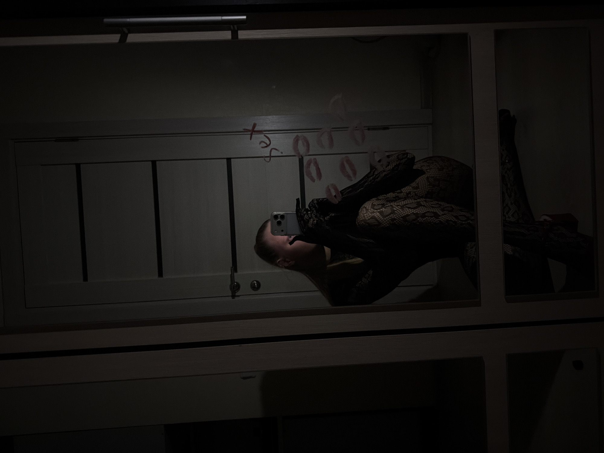 The image displays a dimly lit mirror selfie taken indoors, likely in a bedroom or dressing area in Izhevsk, Russia. A person, partially visible in the reflection, is captured taking the photo with a white smartphone that features multiple rear cameras. The individual, appearing to be a woman with light-colored hair, is wearing dark, patterned attire, possibly a lace or fishnet bodysuit, and is positioned in a reclining or leaning pose against an unseen surface.
The large mirror, which appears to be part of a light-colored wardrobe with paneled doors, is marked with red lipstick. Visible text on the mirror spells "x sex," accompanied by numerous distinct lipstick kiss impressions scattered across the reflective surface. Above the mirror, a metallic horizontal handle is also visible. The low light creates a moody and intimate atmosphere, with only artificial illumination present. Details of the room's furniture, including the paneled doors, and a textured fabric with a snake-skin-like pattern (possibly part of the person's attire or bedding), are discernible through the reflections. The activity depicted is a personal moment of self-photography, framed by the reflective surface and its evocative markings.