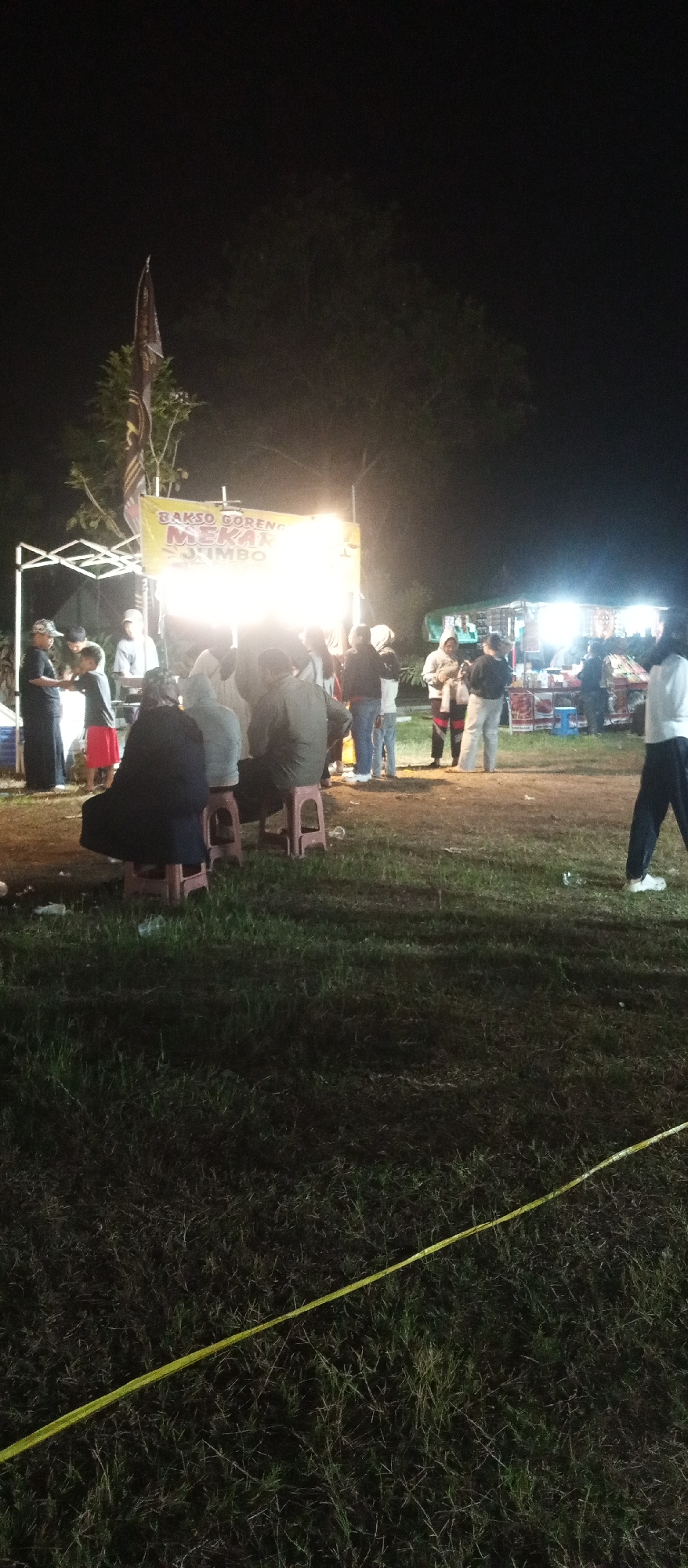 A nighttime scene depicts an outdoor gathering at what appears to be a food stall in Cermo, Indonesia. A large, illuminated banner with text that reads "BAKSO GORENG MEKAR" and "JUMBO" is prominently displayed, indicating a food vendor. Several people are gathered around the stall, some standing and others seated on low stools. The ground is covered in grass. In the foreground, a yellow line is visible, possibly marking a boundary or caution tape. The lighting is artificial, originating from the food stall and some overhead lights. The overall impression is that of a casual community event or market.