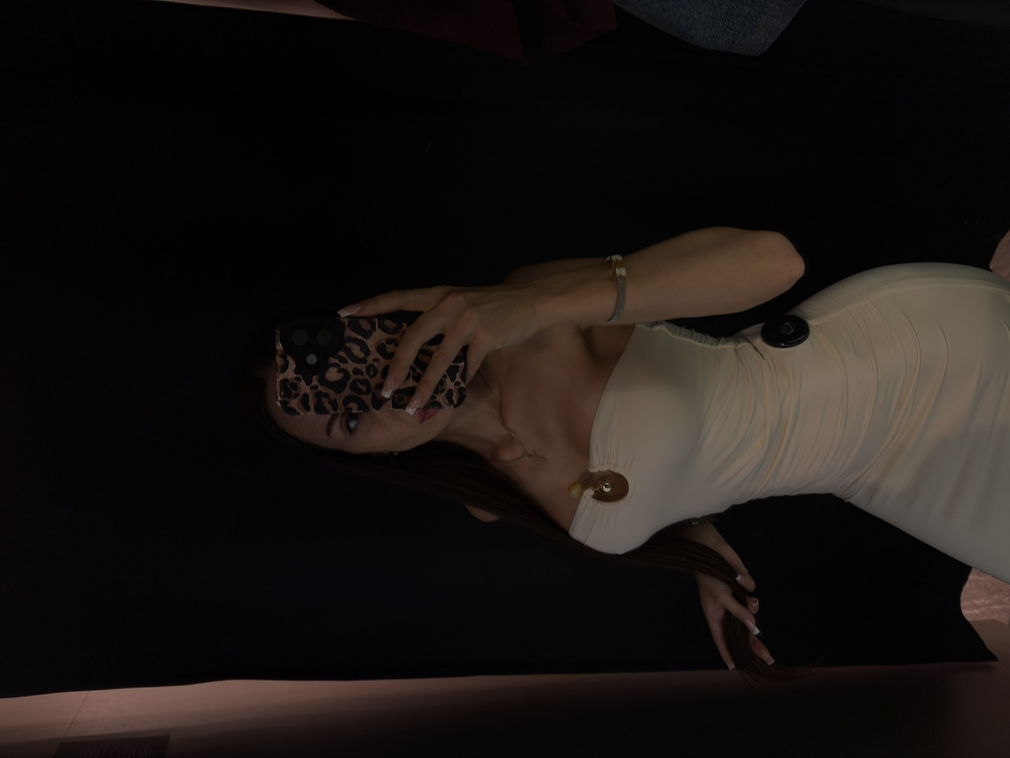 A young woman stands in a dimly lit fitting room, holding a leopard-print phone to partially obscure her face as she takes a selfie. She is wearing a cream-colored, strapless, ruched dress with a gold circular detail at the bust and a black circular embellishment on the side. Her hair is long and dark brown, falling over her shoulders. A delicate necklace with a cross pendant is visible, as is a bracelet on her left wrist. The fitting room appears to be in a clothing store, indicated by the dark curtains and the presence of other clothing items on a rack to the right. The lighting is low, creating shadows and highlighting the woman's form. No specific location in Saint Petersburg, Russia, is identifiable from the image.