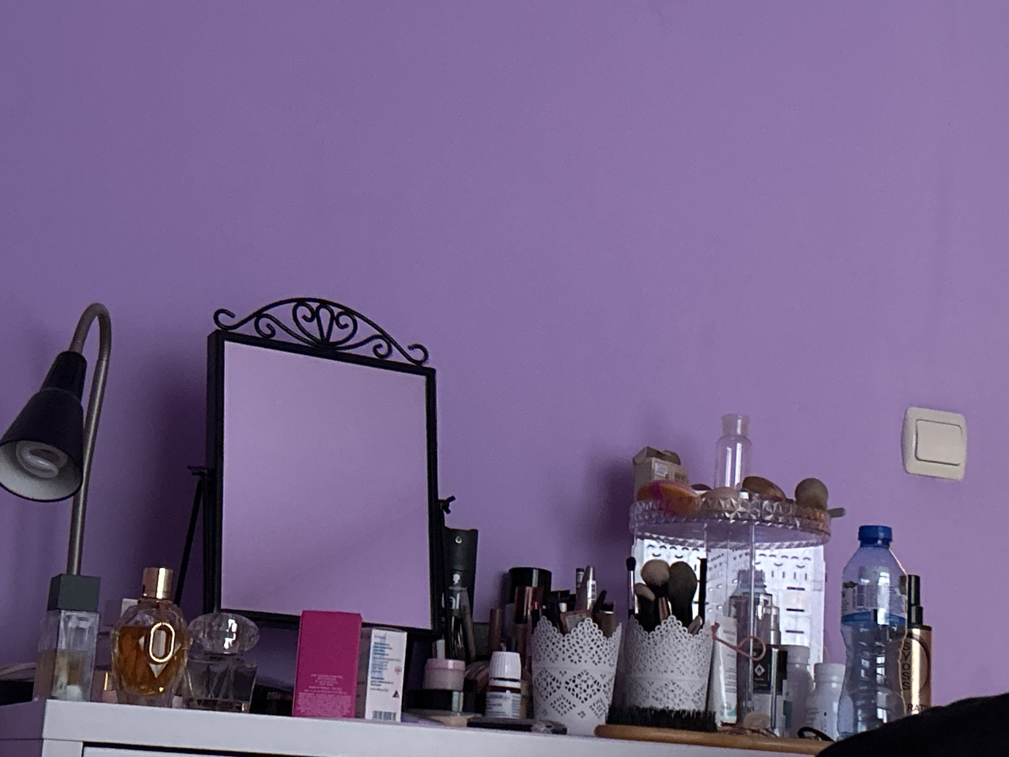 The image depicts a cluttered dressing table in front of a light purple wall. On the left is a black desk lamp and a perfume bottle. A black-framed mirror stands in the center. To the right, there are containers holding makeup brushes, various cosmetics, and a water bottle. A light switch is visible on the wall in the upper right. There is no visible evidence to indicate this scene is in Plovdiv, Bulgaria.