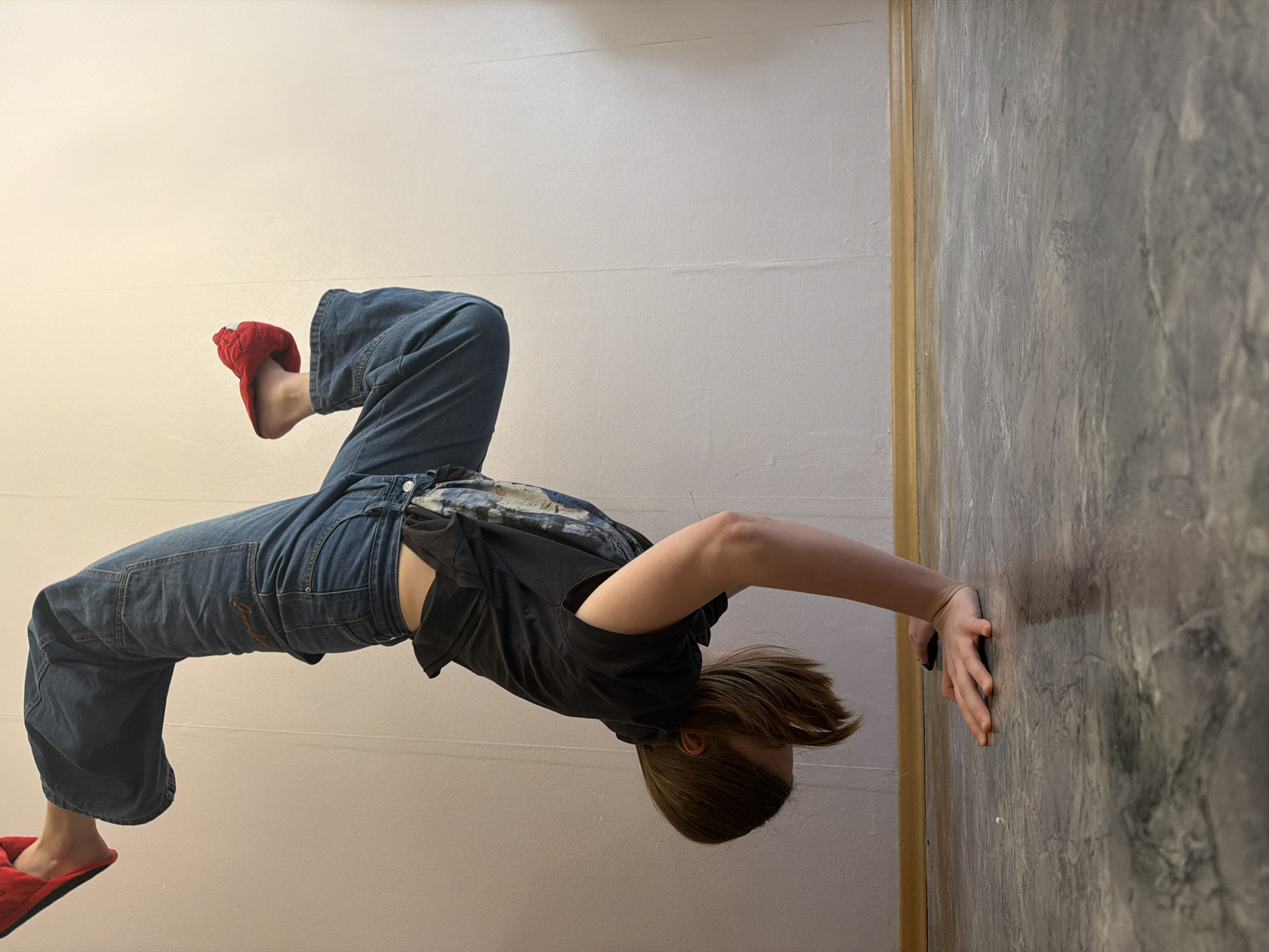 An indoor photograph, taken in Perm, Russia, captures a young person performing an acrobatic pose, possibly a handstand, against a textured grey wall. The image is oriented 90 degrees clockwise, making the person appear to be suspended horizontally.
The central subject is a young individual with medium-length light brown hair, wearing a dark (likely black or dark grey) t-shirt and loose-fitting blue denim pants or overalls. Their feet are clad in distinctive bright red slippers. Their hands are pressed against the textured grey wall, providing support for the pose, while their body is arched with legs extended upwards.
The setting consists of two distinct walls meeting at a corner. One wall, to the left (or bottom due to rotation), is plain and off-white or cream-colored. The other wall, to the right (or top), features a rough, textured grey finish, possibly designed to mimic concrete or distressed plaster. A thin, golden-brown strip delineates the edge where these walls might meet a floor or another architectural element. The lighting appears to be artificial, casting soft shadows, indicating an indoor environment. There is no visible text or discernible emotional expression from the subject. The activity suggests agility or a playful display of physical skill.