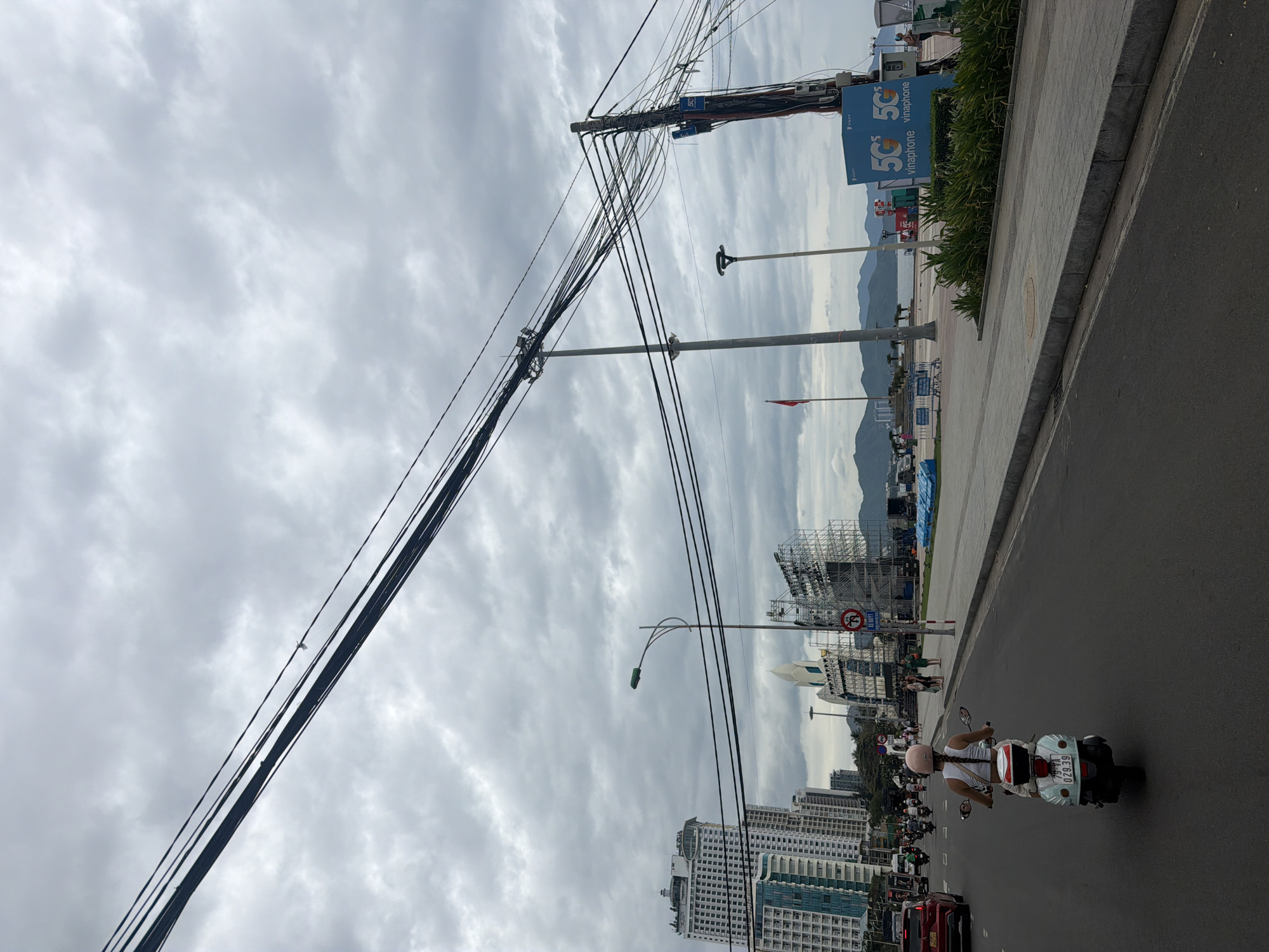 This is a vertical, eye-level shot of a street scene in Nha Trang, Vietnam, under a cloudy sky. In the foreground, a person on a light blue scooter with a white and red storage box on the back is traveling away from the camera on a paved road. The scooter has a license plate reading "79-VA 029.39". The rider wears a light pink helmet and a white tank top with braided hair extending down their back. A red car is visible ahead of the scooter.
The middle ground features a wide, grey paved sidewalk with some greenery. A tall utility pole with a mass of tangled black wires is prominently positioned on the right side of the frame, with a blue "5G VINAPHONE" advertisement attached to it. Further down the street, a large scaffolding structure is visible, along with several tall buildings, including a white high-rise building on the left. A street light with a green lamp is positioned on the left side of the road, in front of the buildings.
In the background, mountains are visible beyond a body of water, suggesting proximity to the coast. A red flag on a pole is also present in the distance. The overall atmosphere is overcast and slightly hazy.