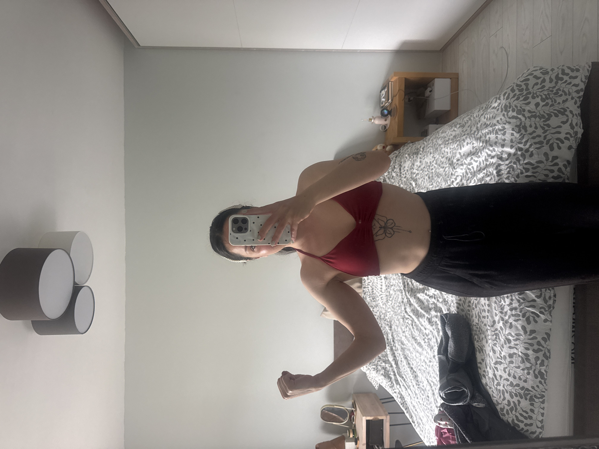 A mirror selfie captures a person in a modern bedroom located in Burgas, Bulgaria. The individual, primarily visible from the waist up, holds a smartphone with a light-colored, polka-dotted case, which partially covers their face. They are wearing a dark red bra or sports top and dark trousers, and their dark hair is pulled back with a light-colored headband. The person is striking a pose, flexing their left arm with a closed fist, potentially showcasing their fitness. Distinctive tattoos are visible: a floral design on the left side of their torso below the breast, and another on their upper right arm.
The bedroom features light-colored walls and what appears to be a light wood floor. A bed, covered with a white duvet adorned with a grey leaf-like pattern, is a prominent feature in the background. To the right of the bed, a wooden bedside table holds various items, including a small light-colored box and a pink/rose gold personal care device. A dark jacket or piece of clothing is draped on the bed. On the left wall, a contemporary light fixture composed of three cylindrical shades (two dark grey, one lighter grey) adds to the decor. The lighting is bright and even, suggesting either natural daylight or ample artificial light. No visible text is present in the image.