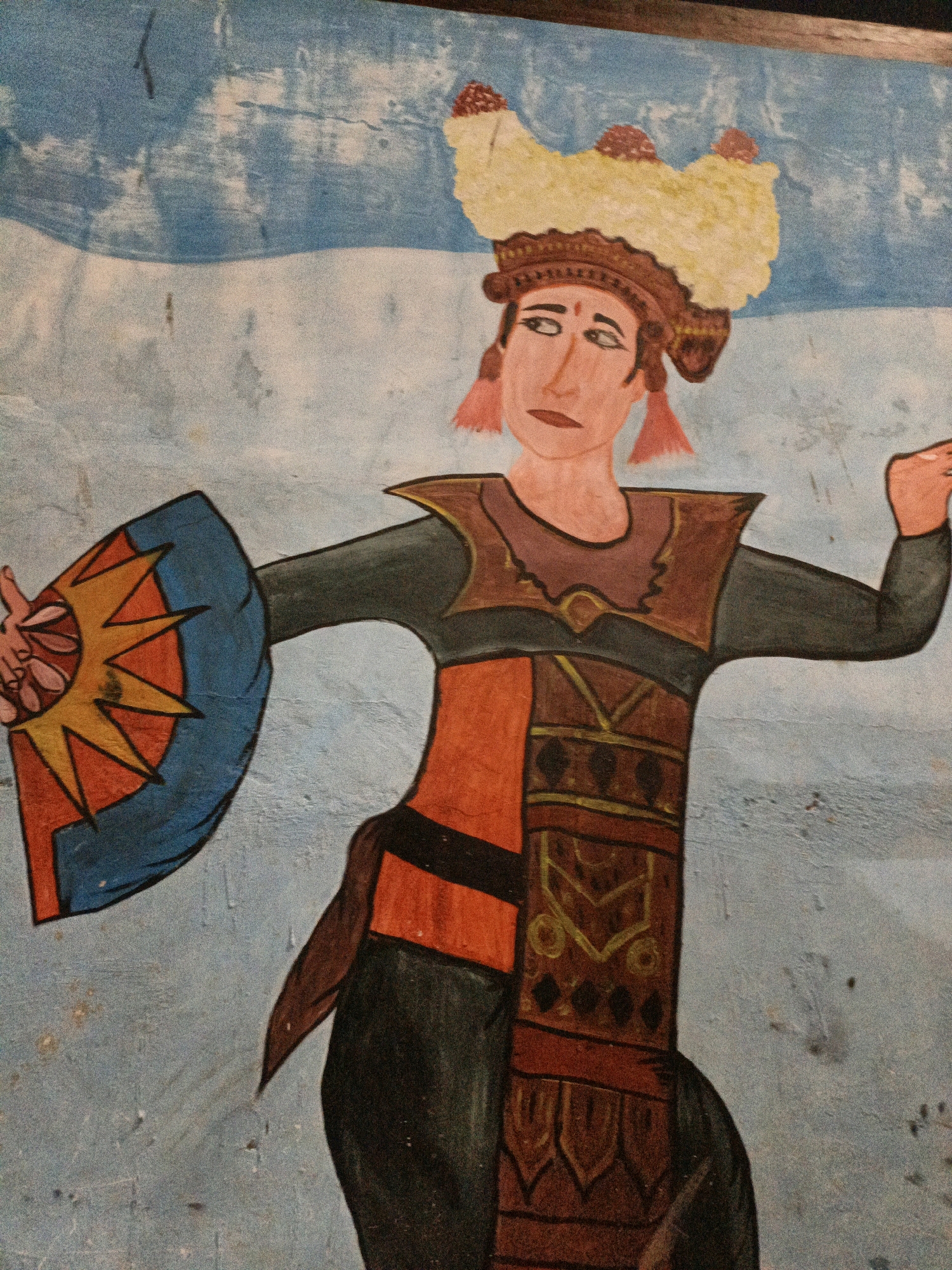 The image is a close-up, medium-angle shot of a painting on a textured, light blue wall. The painting depicts a stylized human figure, likely a dancer, dressed in elaborate traditional attire. The figure has a light-skinned face with dark, almond-shaped eyes and a neutral expression. A large, ornate headdress adorns the figure's head, featuring yellow and red elements. The figure wears a dark green or black long-sleeved garment beneath a layered, ornate costume consisting of brown and red fabrics with decorative patterns. The figure holds a fan-like object in its left hand, which is blue with a yellow and orange sunburst design. The figure's right arm is extended to the side. The painting appears to be somewhat aged, with some scuff marks and discoloration visible on the wall. The location in West Jakarta, Indonesia is not discernible from the image itself.