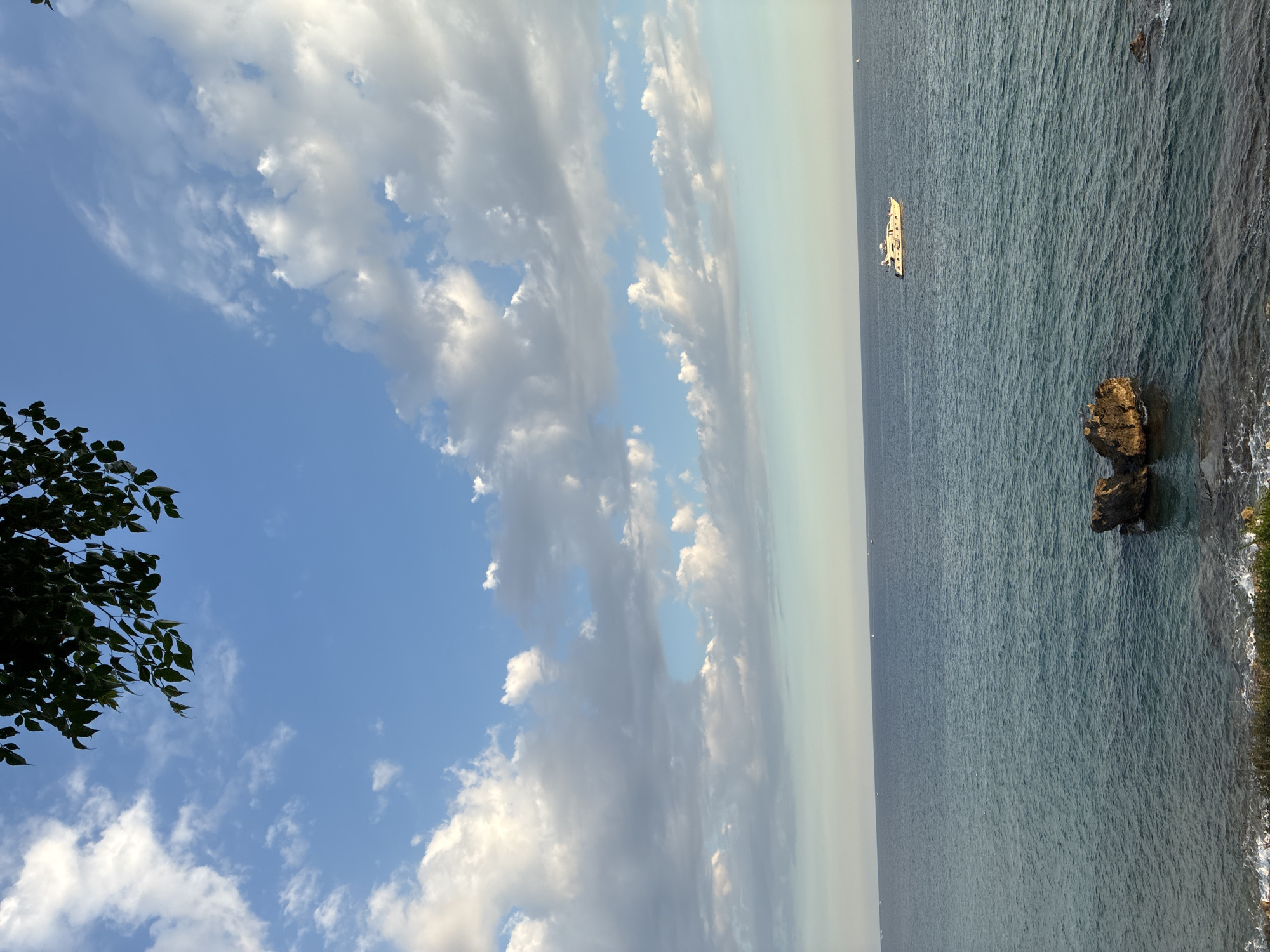 An expansive view of the Mediterranean Sea off the coast of Alanya, Turkey, is captured from a high vantage point. The sky is a bright, clear blue, punctuated by fluffy white clouds that cast subtle shadows on the water below. A prominent white yacht is visible on the horizon, sailing calmly across the blue expanse. In the foreground, dark, jagged rocks emerge from the turquoise water, adding a touch of natural ruggedness to the scene. The coastline is lined with lush green foliage, suggesting a temperate climate. The sun appears to be setting, casting a warm, golden light that highlights the textures of the clouds and the ripples on the water. The overall impression is one of tranquility and natural beauty, typical of the Alanya coastline.