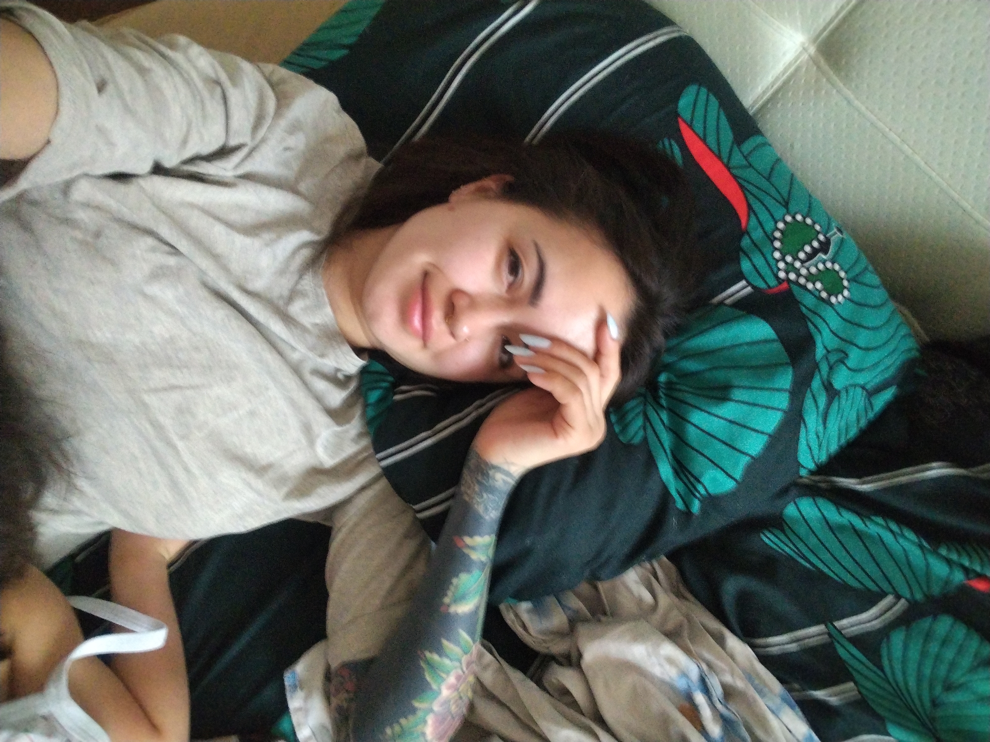 A woman lies on a bed in an interior setting. She has dark hair, fair skin, and visible tattoos on her left arm. She is wearing a gray top. Her right hand is resting near her face. She is lying on a pillow with a green and black patterned design. The bed has a white tufted headboard. Part of another person with dark curly hair and a white tank top is visible in the lower left corner of the image. The location in the city of Noviy, Russia, is not discernible from the image.