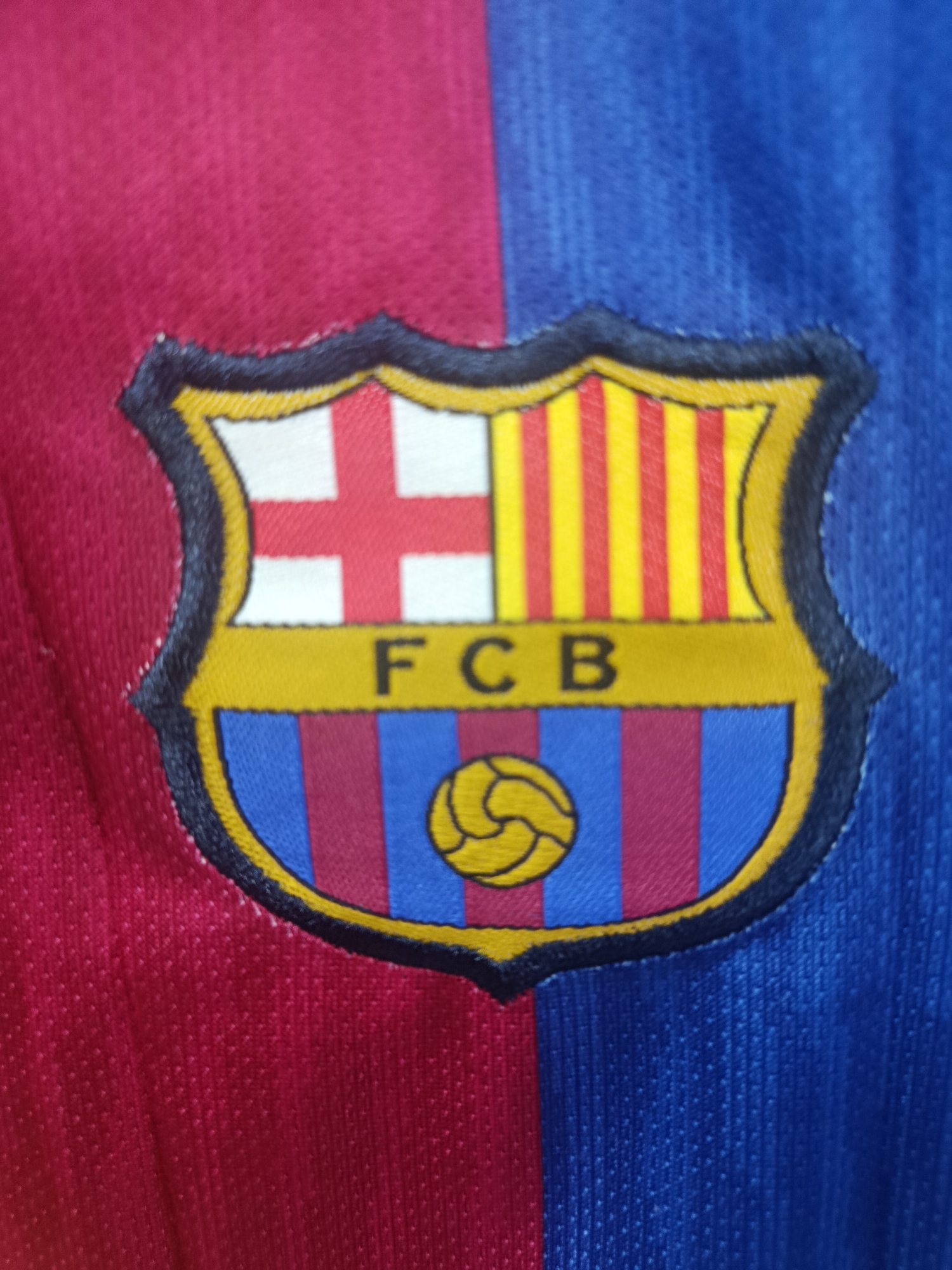 The image is a close-up, high-angle shot of a F.C. Barcelona crest embroidered on a red and blue striped fabric, likely a jersey. The crest is a shield with a red and white cross on the left side, and yellow and red vertical stripes on the top right. Below this is a yellow band with the letters "FCB" in black. The bottom of the shield features blue and maroon vertical stripes, with a yellow soccer ball in the center. The fabric appears to be a synthetic material, possibly polyester, with a textured surface. The crest is bordered by a black and yellow embroidered edge. The background is the red and blue fabric of the garment. There are no people or any indication of a specific city, including Sertolovo, Russia, in the image.