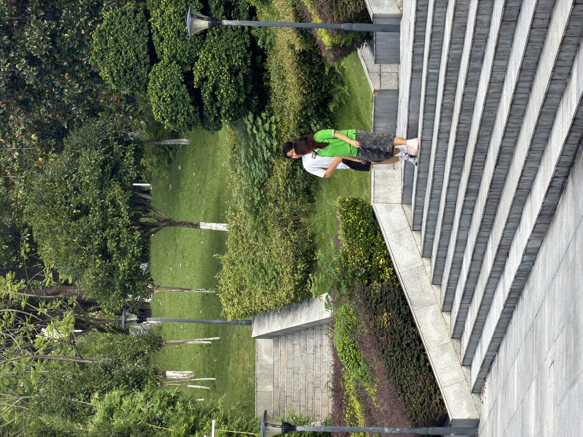 Two young individuals, a male wearing a white t-shirt and dark shorts and a female in a green polo shirt and camouflage shorts, are standing on a wide, gray concrete staircase. They are positioned on one of the middle steps of a long flight of stairs that ascends a grassy incline. The stairs are made of stone or concrete blocks and are flanked by planters filled with dark red and green bushes. Beyond the staircase and manicured bushes, the landscape features lush green grass dotted with trees and more dense foliage. Several dark gray lampposts are visible, one on the lower left and another further up on the right, both with a dark, rounded top. In the background, mature trees and hedges create a dense, green environment. The overall setting appears to be a public park or outdoor recreational area in the city of Jiangjin, China.