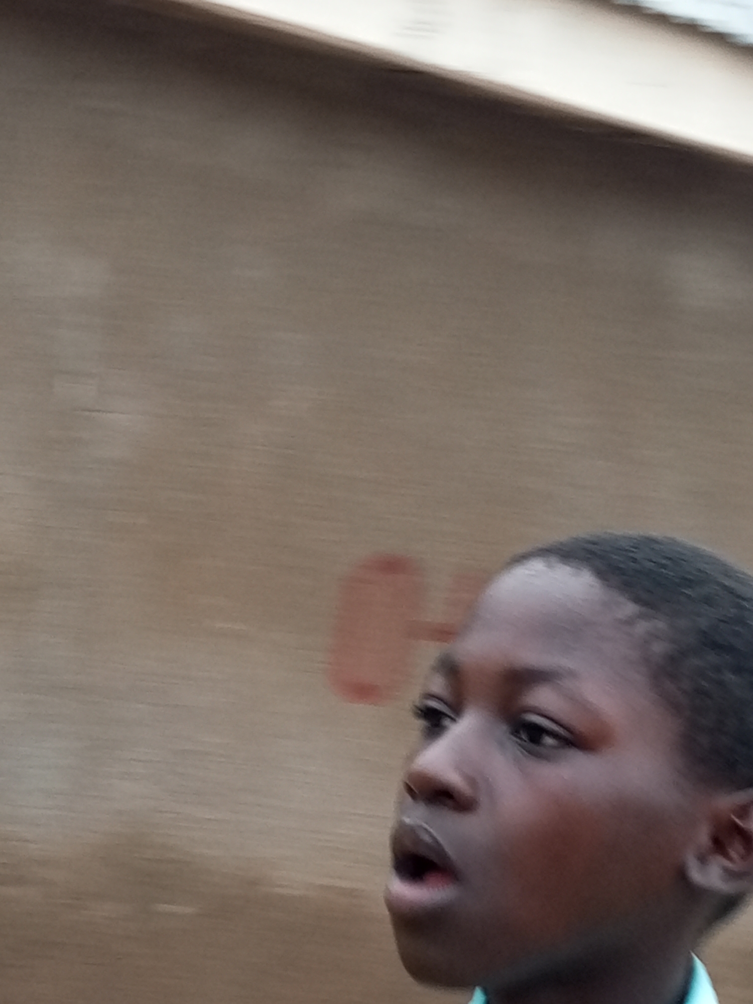 The image shows a young Black boy looking to his left, with his mouth slightly open. He is wearing a light blue collar shirt. The background is a textured, light brown wall. There is a faint, reddish-orange circular mark on the wall to the left of the boy. The lighting suggests it is daytime. The scene appears to be outdoors. The location in the city of Tempure, Nigeria, is not discernible from the image.