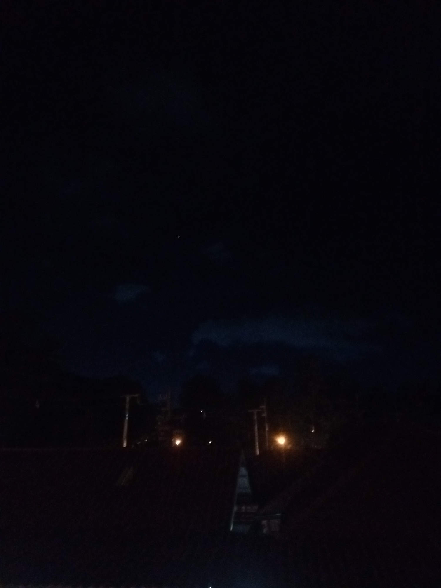 The image depicts a night scene in Baan Chaloklum, Thailand. The sky is dark, with visible clouds and a single bright star. In the foreground, there is a dimly lit rooftop. Beyond the roof, there are trees and utility poles. The street lights cast a faint glow in the distance. The scene is dark, with limited details visible due to low lighting.