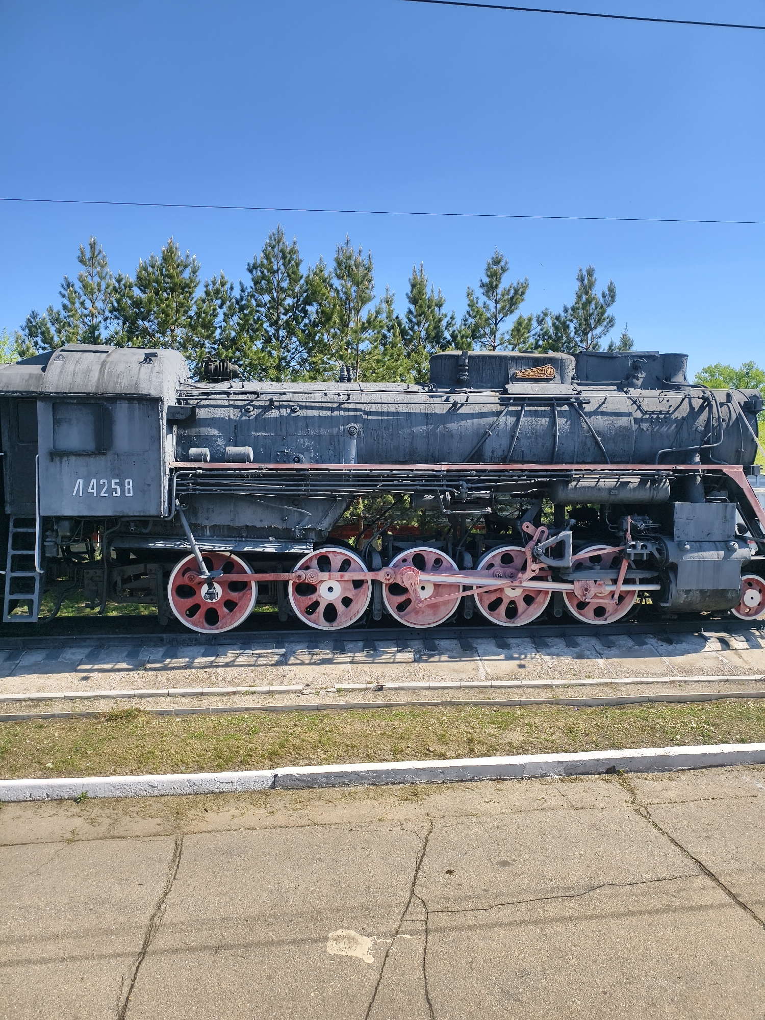 The image depicts a gray steam locomotive, numbered "Л 4258", displayed outdoors. The locomotive has red wheels and sits on rails embedded in concrete slabs. A grassy strip separates the rails from a cracked asphalt surface. In the background are several coniferous trees against a clear blue sky. The photo was taken in Shimanovsk, Russia. No people are visible.
