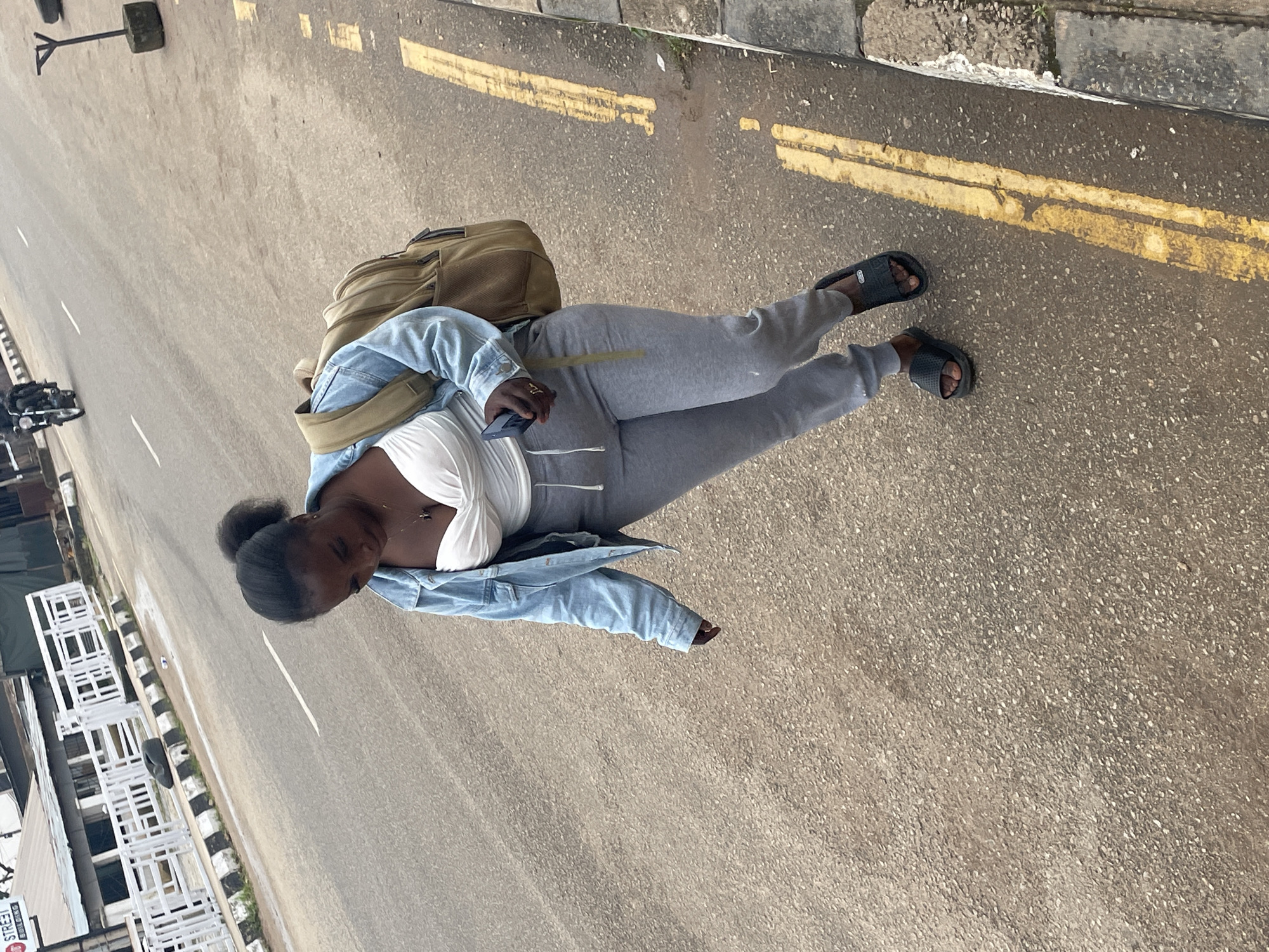 A young Black woman with her hair pulled back in a ponytail is walking on a paved road. She is wearing a light blue denim jacket, a white top, grey sweatpants, and black slides. She has a tan backpack on her shoulders and is holding a black smartphone in her right hand. The woman is looking down at the phone. Yellow lines are painted on the road to mark lanes. In the background, there is a white railing along the side of the road, a motorcycle with two riders, and a building with signage that reads "STREET BUILDING." The environment appears to be a city street in Jos, Nigeria.