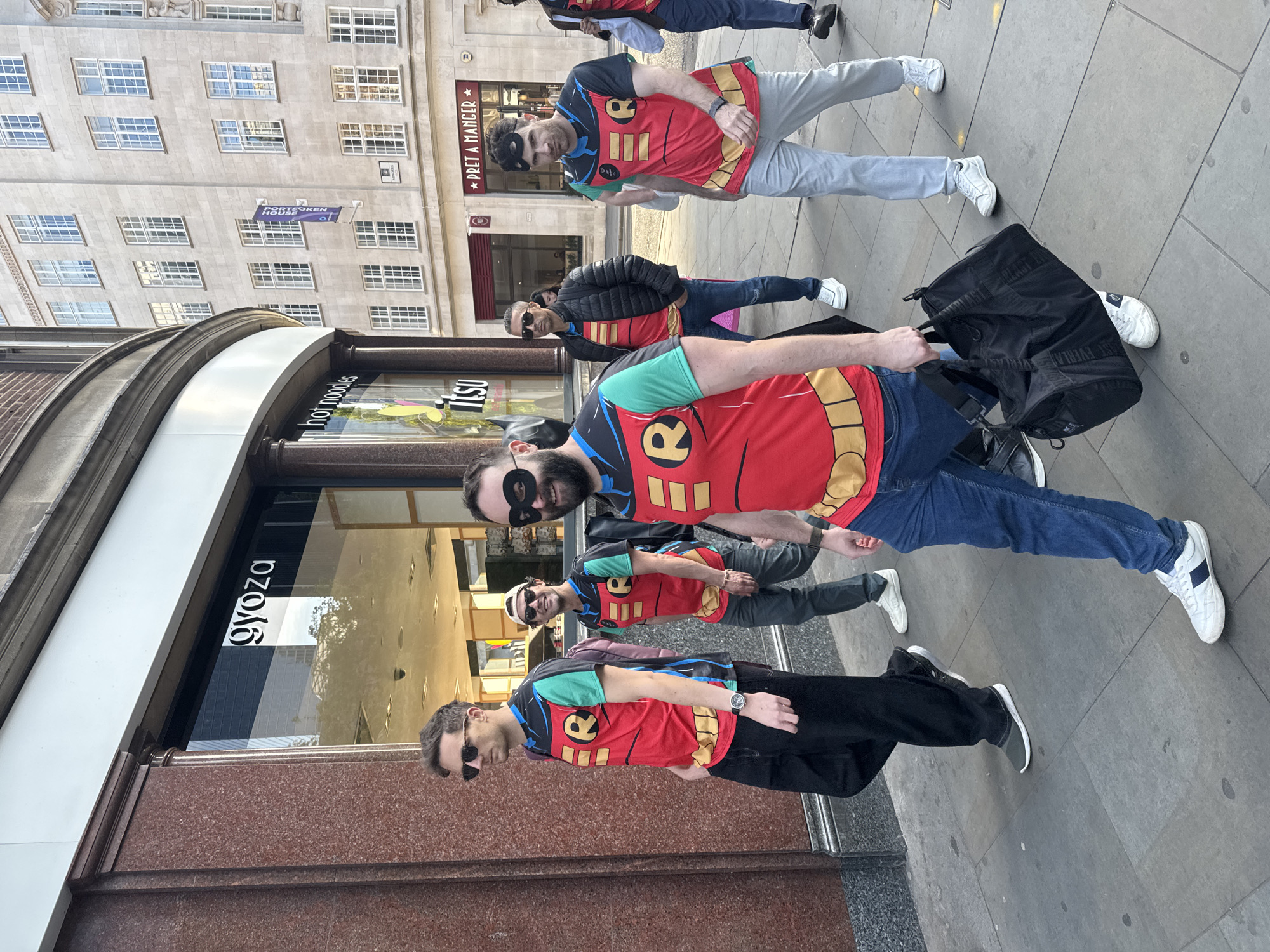 A group of five men dressed in Robin superhero costumes are walking on a city sidewalk, with "gyoza" signage visible in the background, and a "PRET A MANGER" sign on the building across the street. Four of the men are wearing Robin-themed T-shirts with an "R" emblem, while one man has a black jacket over his costume. Some of the men have black eye masks. The man closest to the camera is carrying a large black duffel bag. The architectural style of the buildings suggests an urban setting in an unknown city. The sidewalk is composed of grey paving stones.
