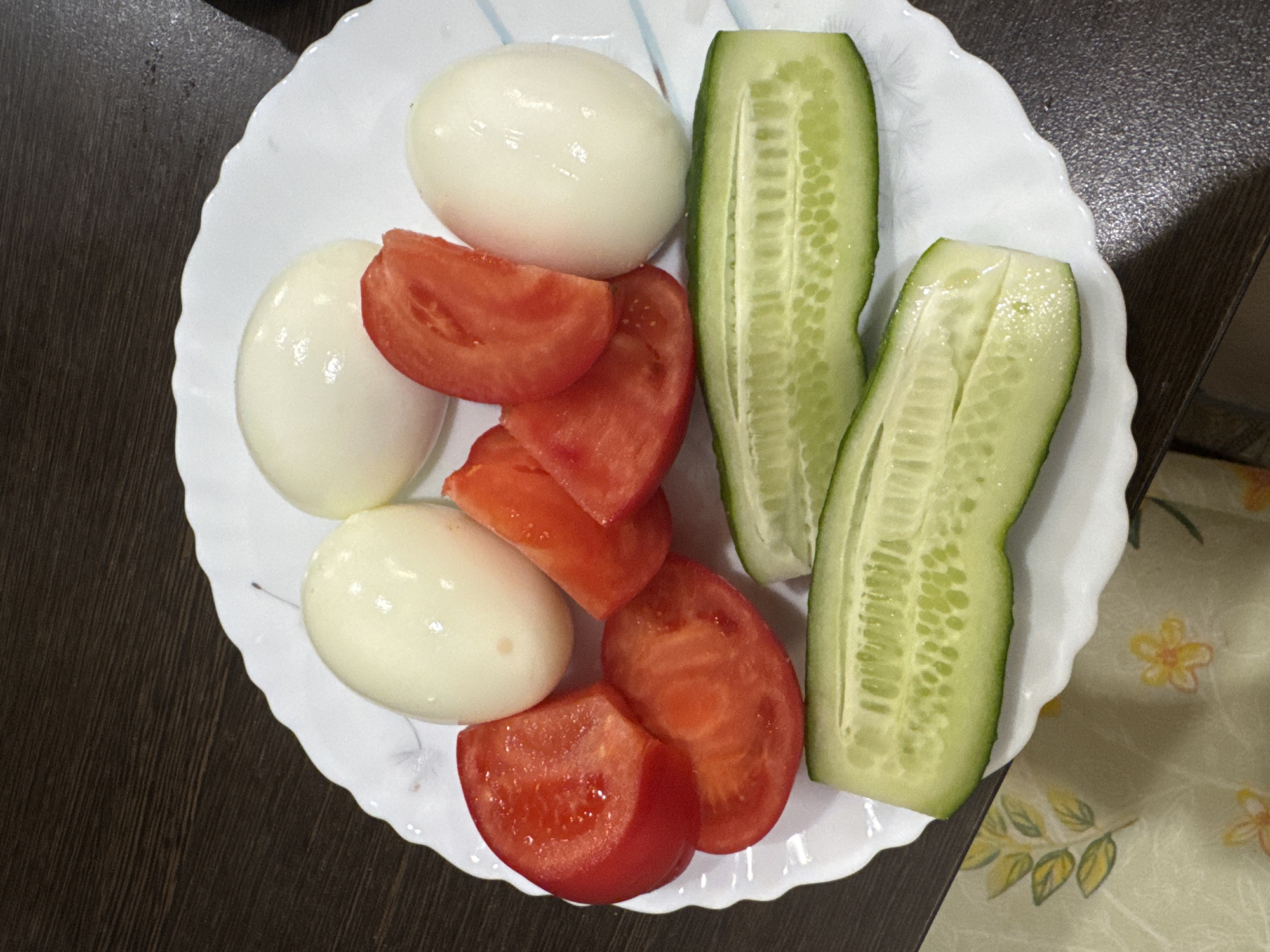 On a white, scalloped-edged plate, there is a selection of food items. There are three peeled, hard-boiled eggs, sliced tomatoes arranged in a stack, and two halves of cucumber cut lengthwise. The plate rests partially on a dark, wooden surface and partially on a light-colored surface with a floral pattern. The location is the city of Sochi, Russia.