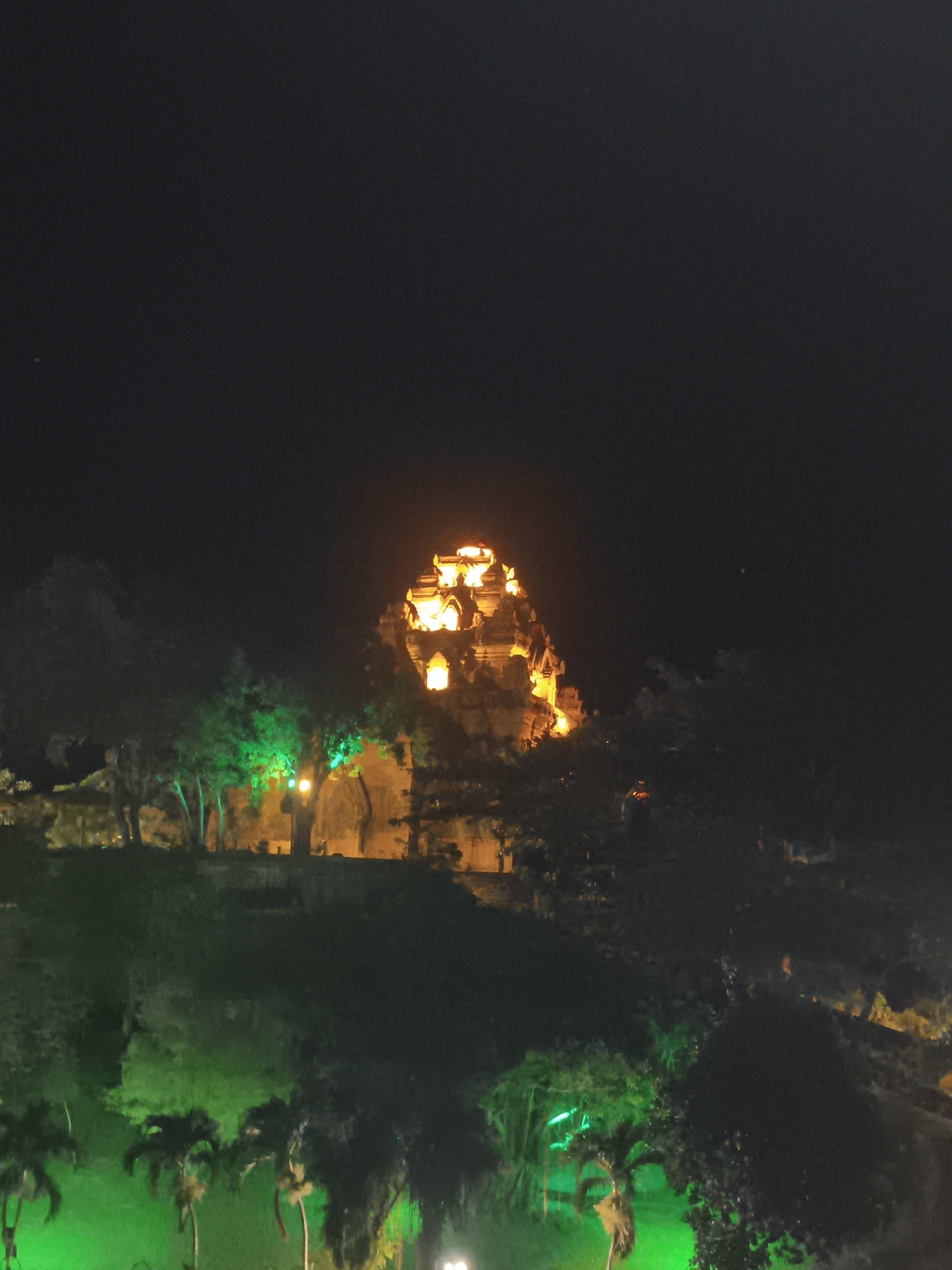 The image captures a nighttime view of a brightly lit temple structure nestled within a lush, dark landscape. The temple, characterized by its tiered, pyramid-like architecture, glows with warm, golden illumination, highlighting intricate details and openings. Surrounding the temple, trees are visible, some of which are illuminated with vibrant green lights, creating a striking contrast against the dark sky and foliage. The foreground shows a sloping, dark green area, possibly a hill or park, with palm trees visible at the bottom left, also bathed in green light. The overall scene suggests a well-maintained park or tourist attraction in Nha Trang Ward, Vietnam, illuminated for evening viewing.