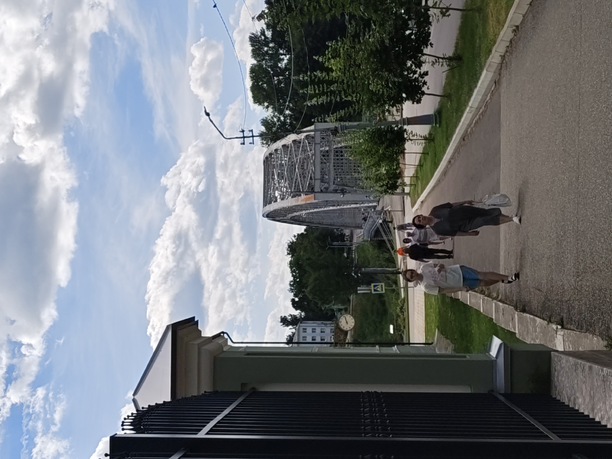 A full shot shows a street scene in Borovichi, Russia, on a cloudy day. In the foreground, a black metal fence runs along the left side of the frame, bordering a light green building with a white roof. A large clock is visible on the side of the building. A paved walkway runs diagonally from the foreground towards the background, with several people walking on it.
On the walkway in the mid-ground, a young person in a white t-shirt and blue shorts walks towards the camera. To their right, a woman in a dark gray dress and white sneakers walks away from the camera, carrying a white bag. Several other people are visible further down the walkway.
In the background, a large, arched steel bridge spans across a body of water. Trees and buildings are visible on the other side of the bridge, and the sky is filled with white, fluffy clouds. A lamppost stands to the right of the bridge. The overall impression is a peaceful street scene in a Russian city.