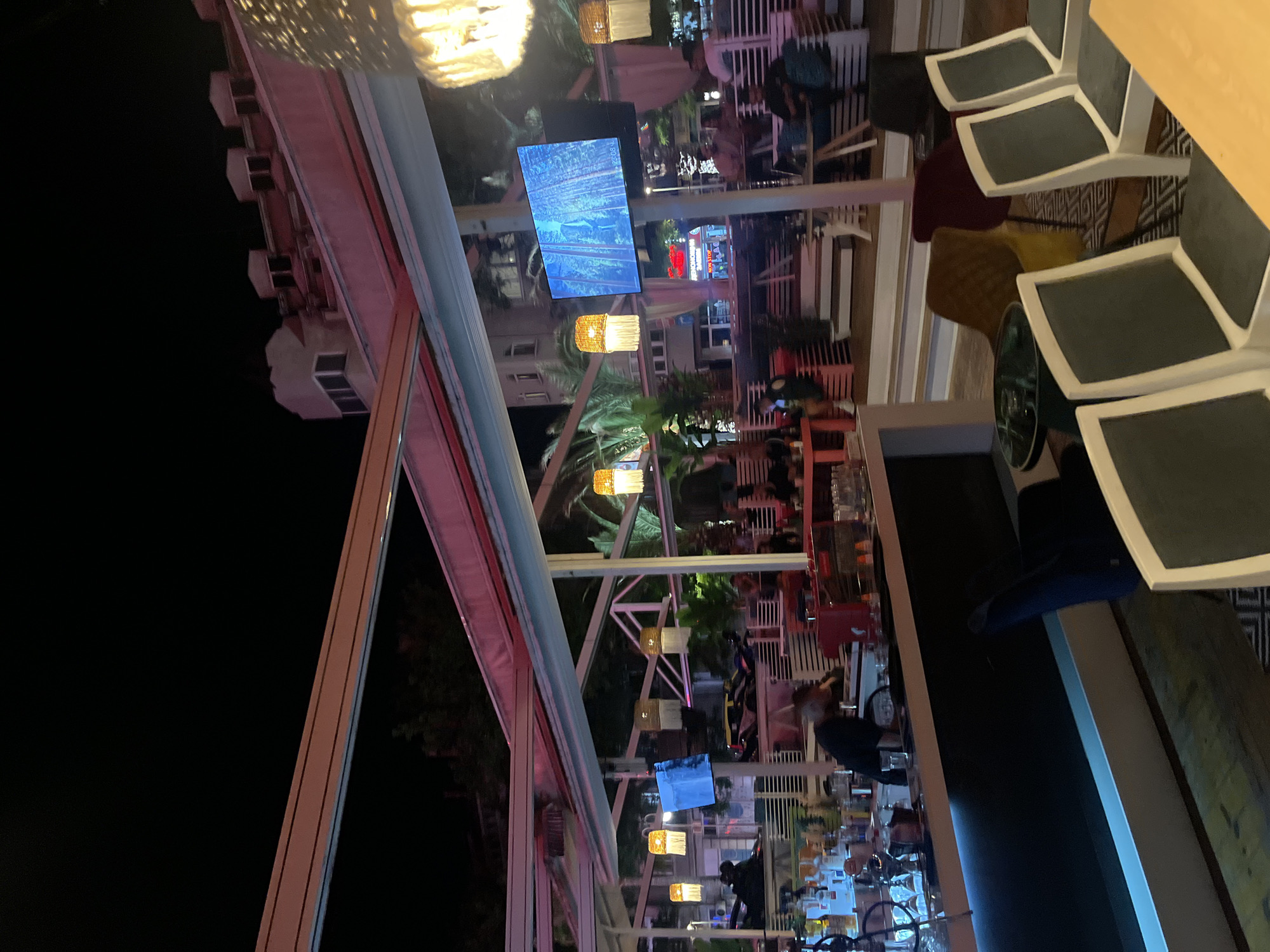 The image displays an outdoor or semi-enclosed bar and lounge area at night, identified as "Neponosim Zamysel" (Intolerable Design) located in Plovdiv, Bulgaria. A white metal framework, illuminated with reddish-pink light, supports an overhead canopy. The area features multiple tiers of white slatted benches and various tables with diverse seating options, including chairs with white frames and dark grey cushions, a dark blue velvet armchair, a yellow upholstered chair, and a maroon upholstered chair.
In the lower-left foreground, a modern bar counter with a dark surface and a blue LED strip along its base is visible. Behind the bar, a male bartender in a dark shirt is engaged in work, surrounded by numerous bottles, glassware, and a red espresso machine. A shisha pipe rests on the bar top.
Several decorative hanging lights with wicker or rattan-style shades and white tassels emit a warm, orange glow. Two large flat-screen televisions are mounted on poles, displaying dynamic blue-toned images; the right screen shows the time "23:08." Green foliage and palm trees are integrated into the design of the lounge area.
Multiple patrons are present, seated at tables or on benches, engaged in social interactions. In the background, a multi-story building with a light facade and prominent dormer windows is visible, alongside other urban structures and trees. Faint vehicle lights suggest an adjacent street. The overall scene is brightly illuminated by artificial lighting, creating a vibrant atmosphere.
