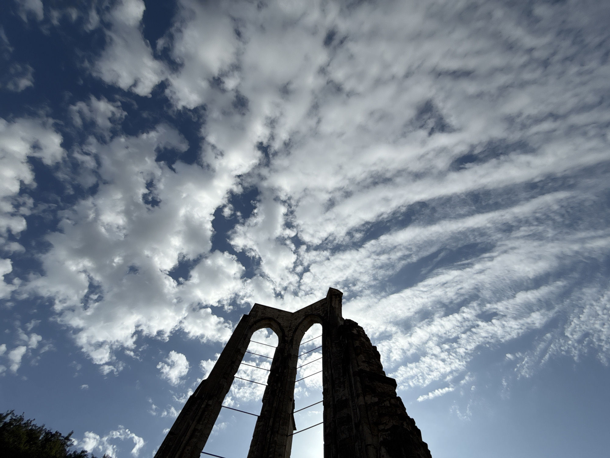 An upward-facing shot shows the sky through the arched ruins of the former Walkenried Abbey in Walkenried, Germany. The clouds are a mixture of fluffy white cumulus and wispy cirrus formations, stretching across a deep blue sky. The stone arches of the ruins are silhouetted against the bright sky, with thin metal supports visible within the arches. The overall impression is one of grandeur and decay, with the natural beauty of the sky contrasting with the man-made structure.